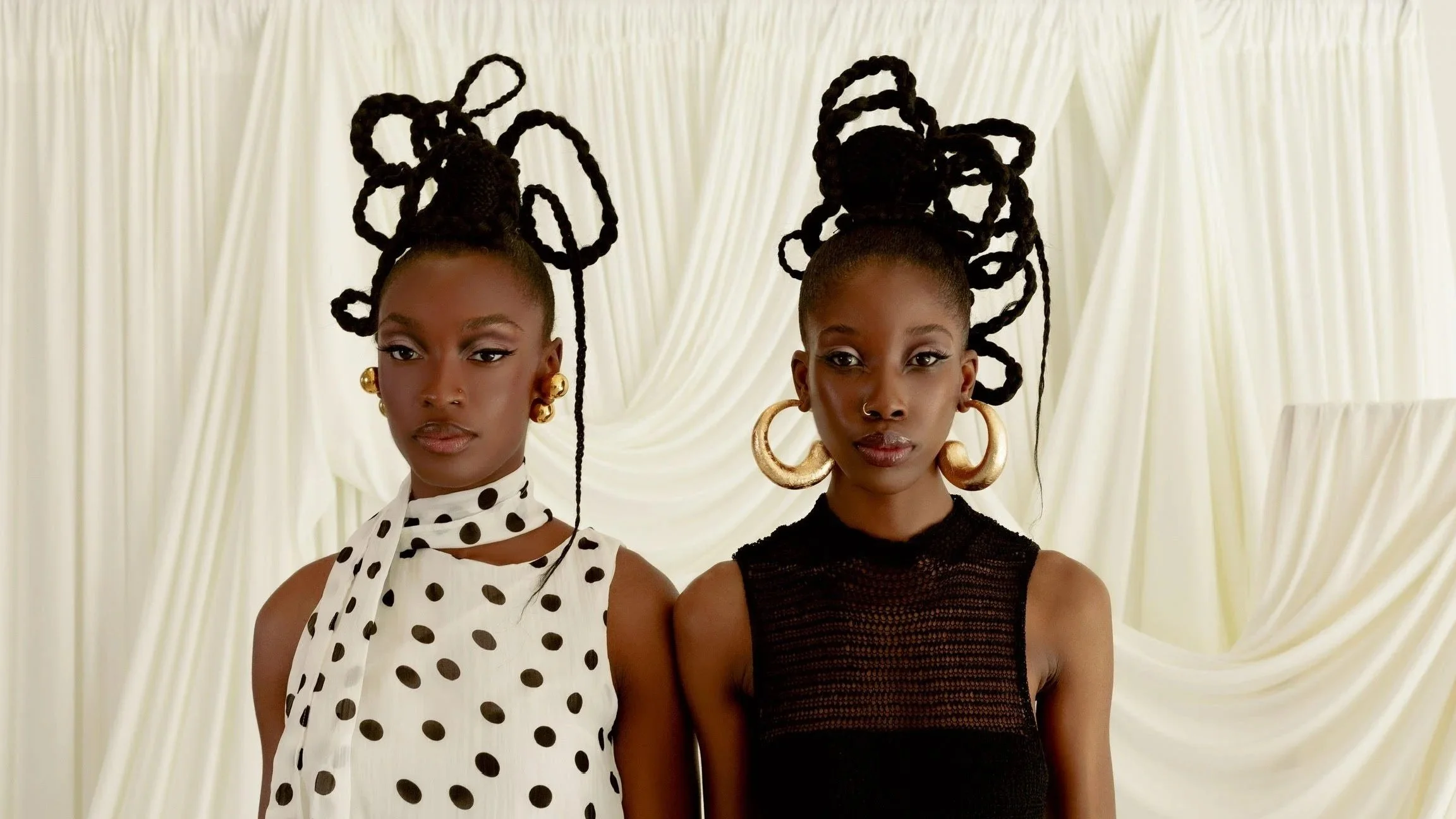 Two women with elaborate braided hairstyles and large gold hoop earrings, standing in front of cream-colored curtains.