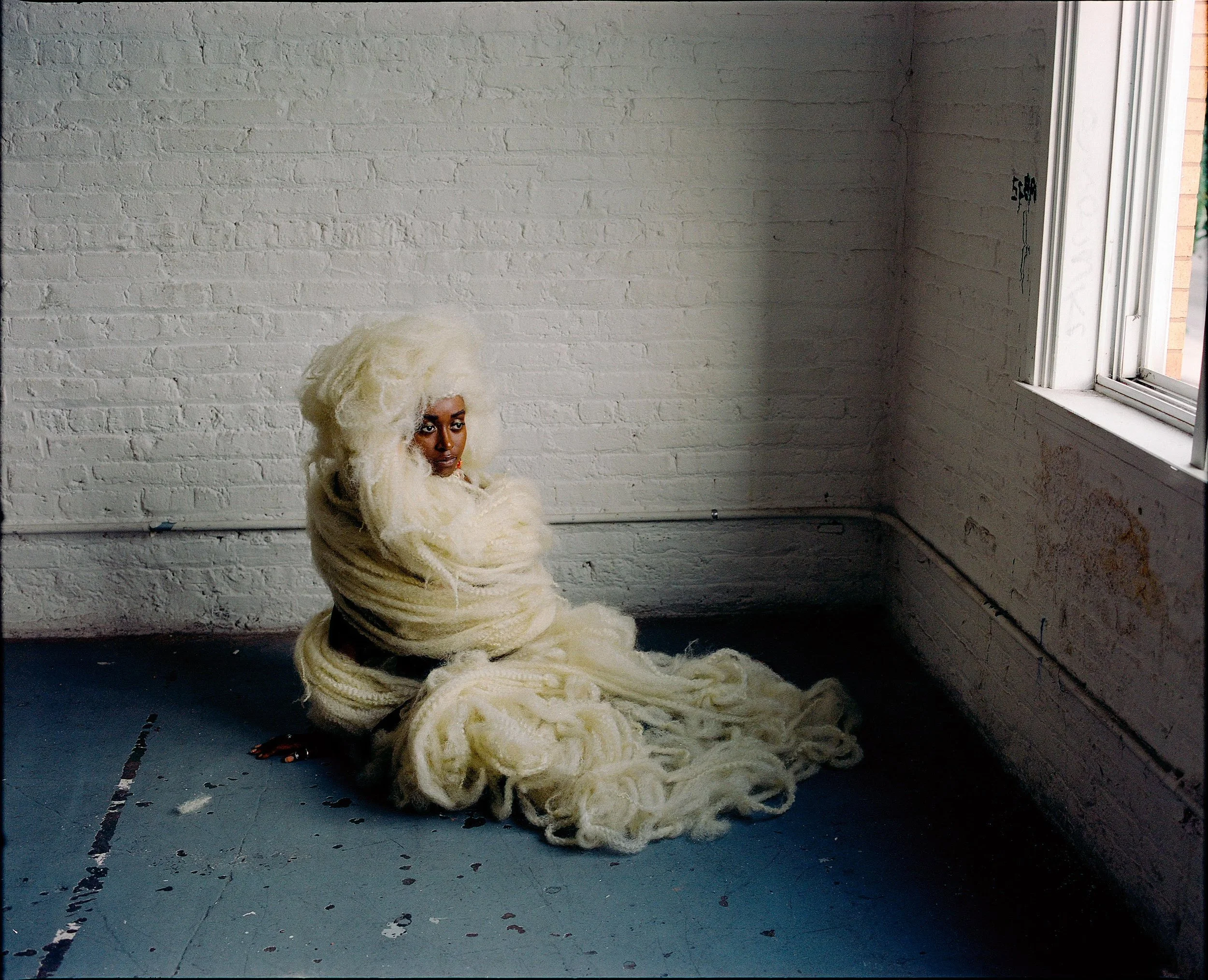 A woman sitting against a white brick wall with a large pile of cream-colored yarn in her lap, near an open window, in a minimalist, industrial-style room.