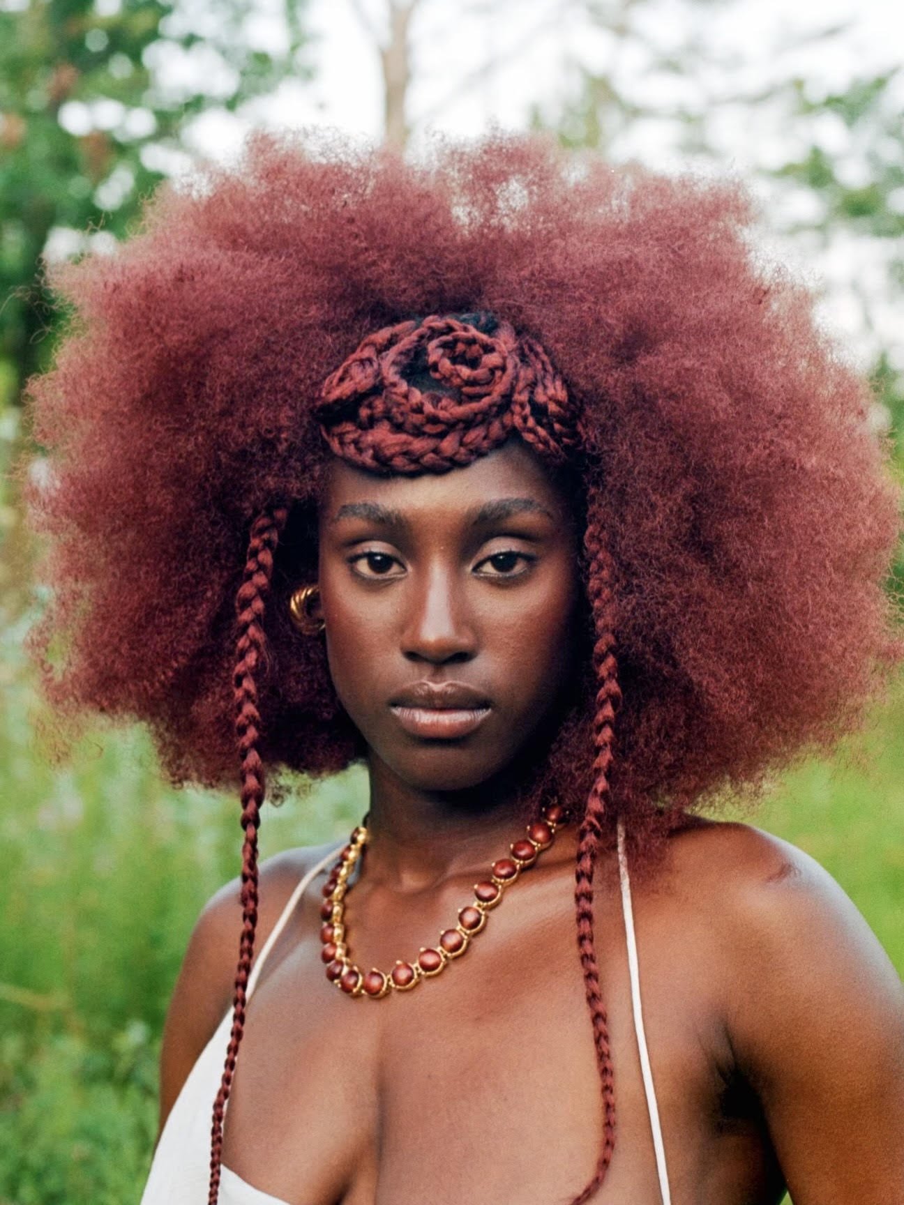 A woman with large, voluminous red curly hair styled with braids and a headwrap, standing outdoors with a blurred green background.