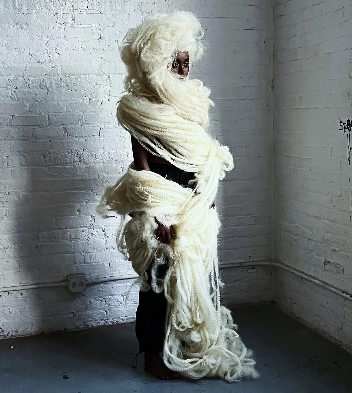 A person standing against a white brick wall, wrapped in large, fluffy, cream-colored fabric or wool, with only their face and feet visible.
