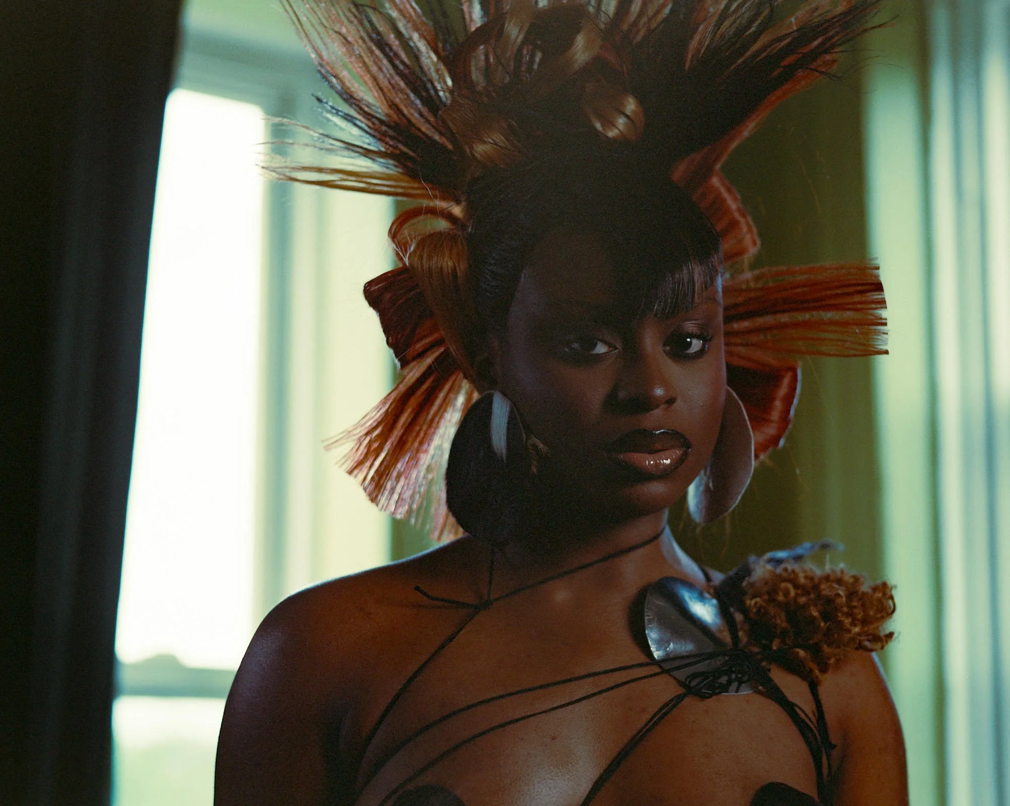 A woman with dark skin and curly hair, wearing black clothing and jewelry, holding a microphone and a plate of food, standing near a window with green curtains.