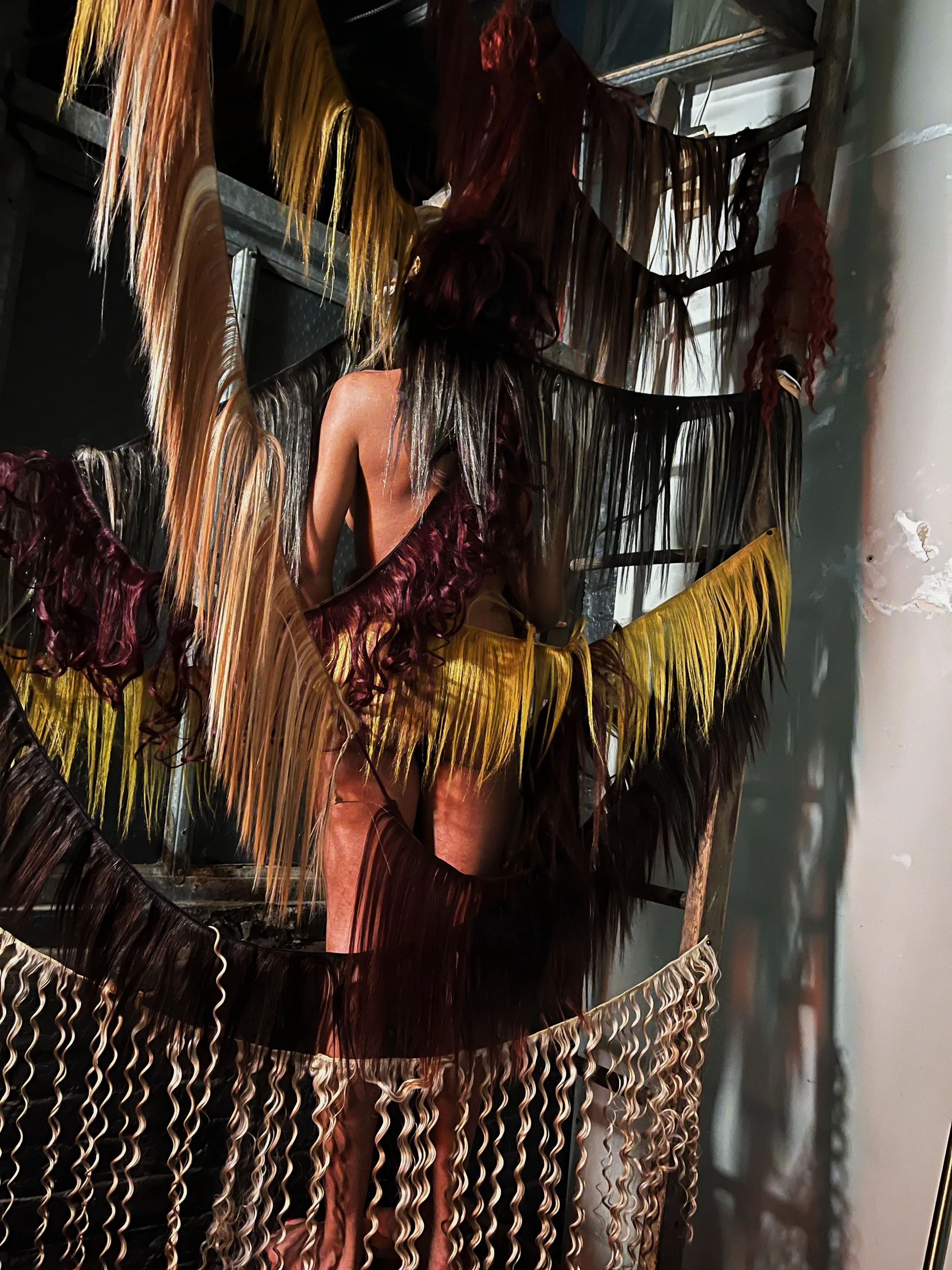 Multiple hair extensions hanging on a rack, with a mannequin wearing a wig behind it. The extensions are in various colors including yellow, red, black, and brown, and some are curly while others are straight.