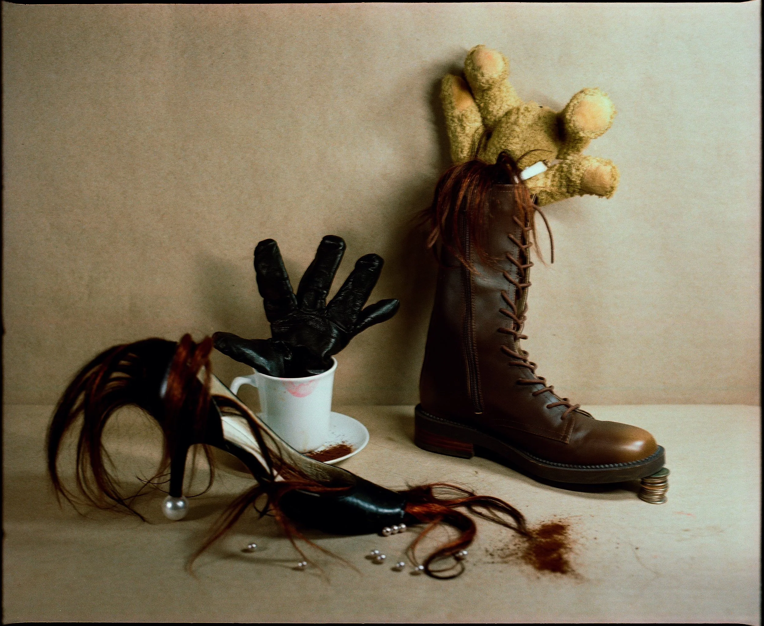 A display of various footwear including a brown leather boot, several pairs of shoes, and a pair of black leather gloves with a white ceramic cup containing a brown hair extension, placed against a beige wall.
