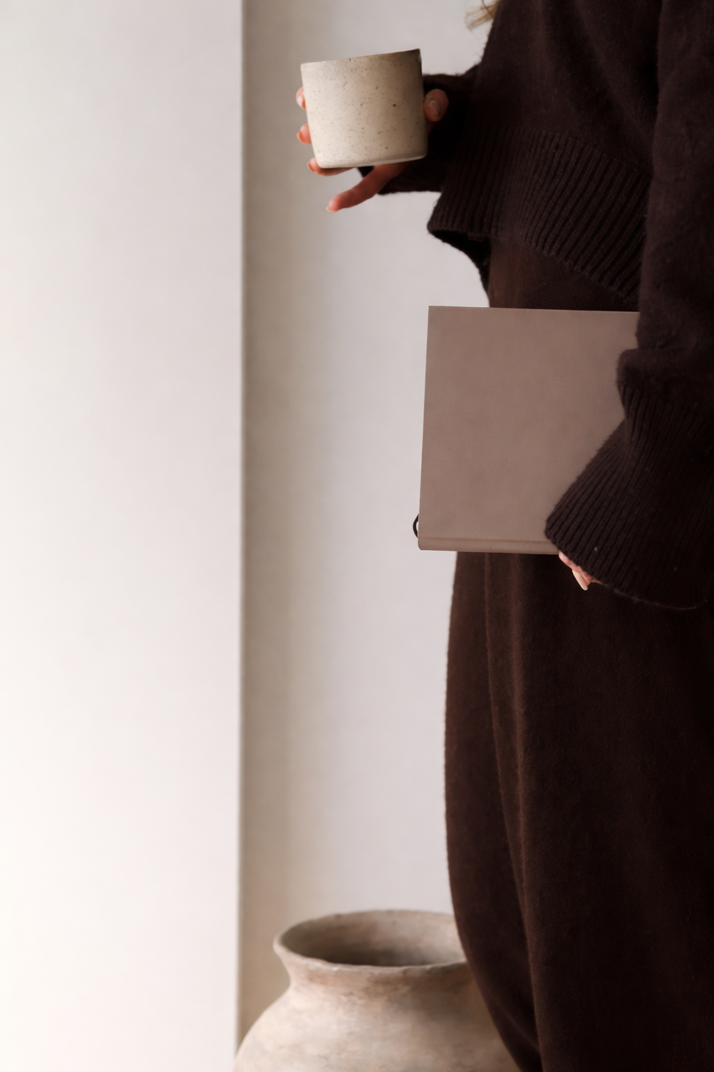 Personne tenant une tasse en céramique et un carnet ou tablette, habillée d'un pull ou manteau foncé, debout à côté d'un pot en terre cuite sur un fond neutre.