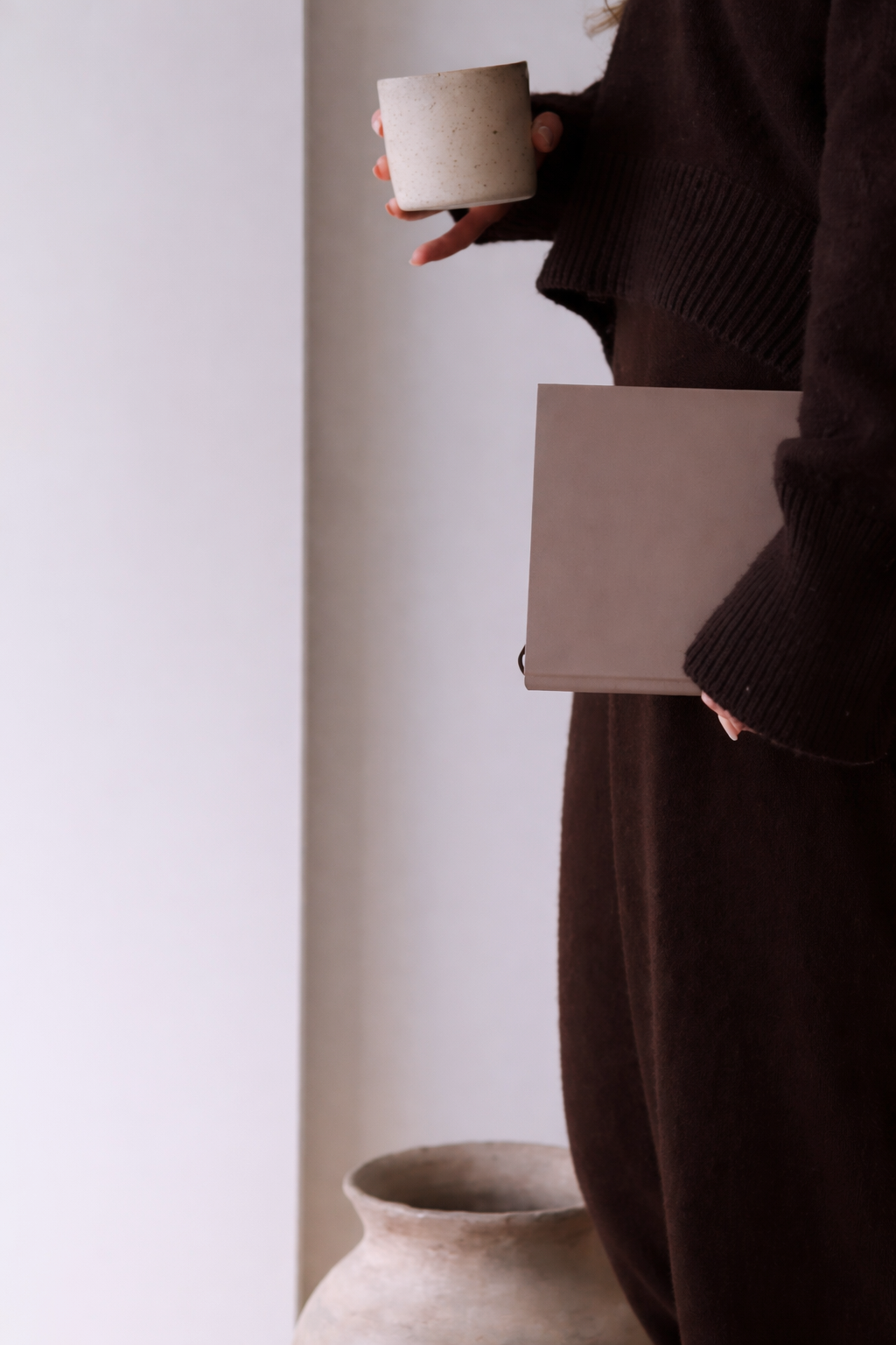 Personne tenant une tasse et un carnet, portant un vêtement sombre, dans un environnement minimaliste avec un pot en céramique au sol.