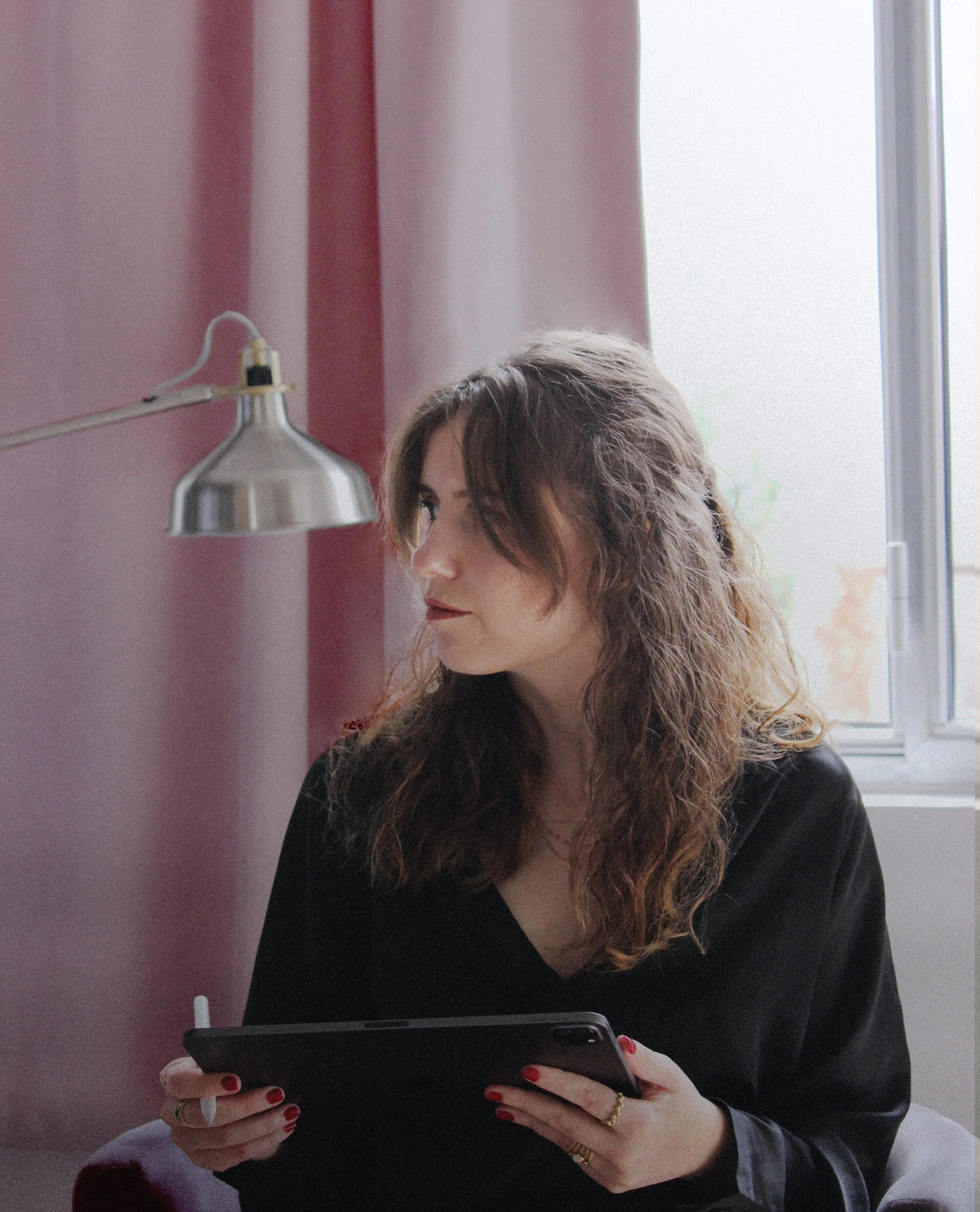 Femme avec cheveux longs et bouclés, portant un vêtement noir, tenant une tablette ou un stylet, assise près d'une fenêtre avec un rideau rose clair, lampe argentée en arrière-plan.