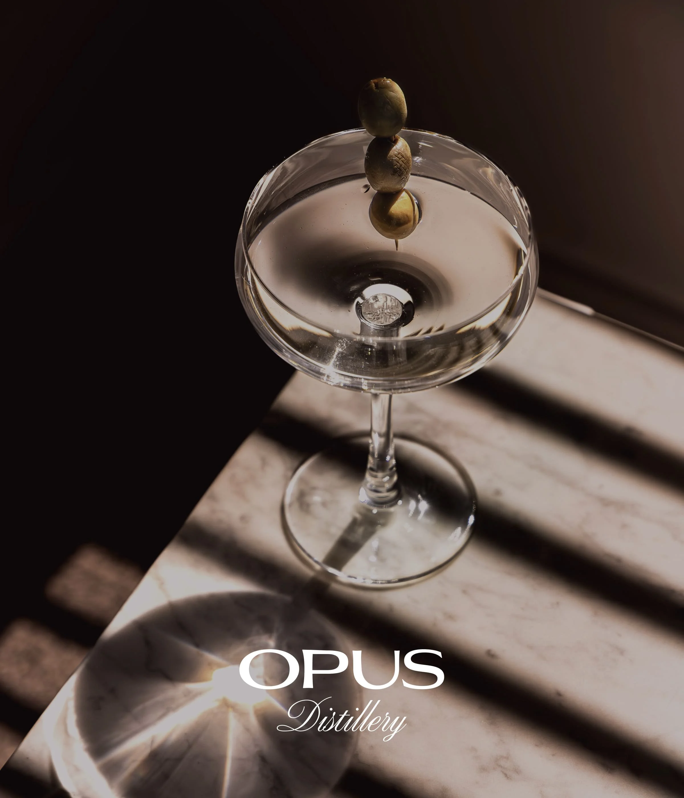 Verre à cocktail avec trois olives empilées, reflet dans le liquide, inscription 'OPUS Distillery' en dessous, plan rapproché sur une surface en marbre, arrière-plan sombre.