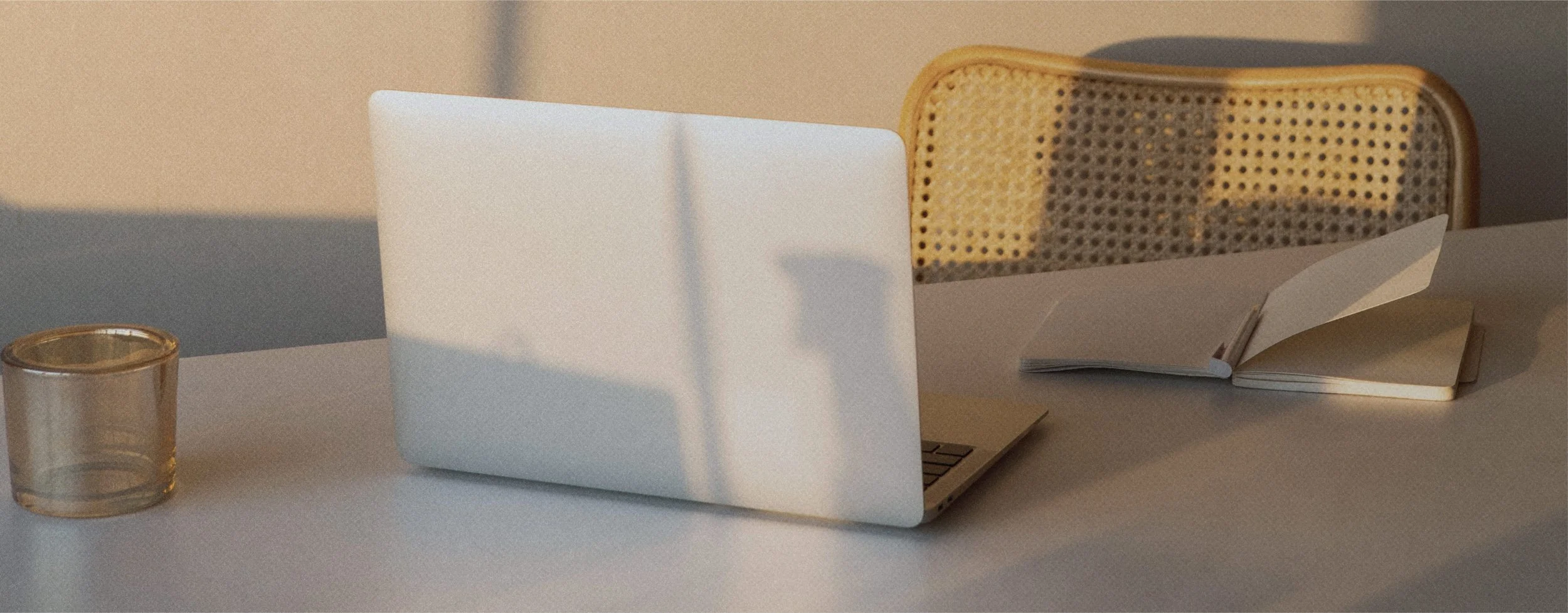Bureau avec un ordinateur portable, un carnet ouvert, un verre transparent, et une chaise en rotin.