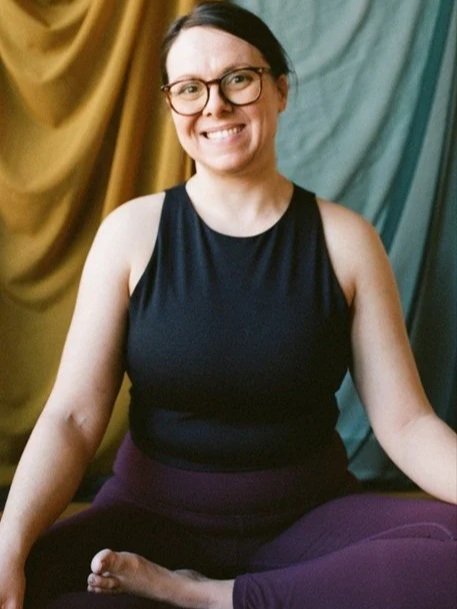 A woman sitting cross-legged, smiling, with glasses, wearing a black sleeveless top, in front of a backdrop with multicolored curtains.