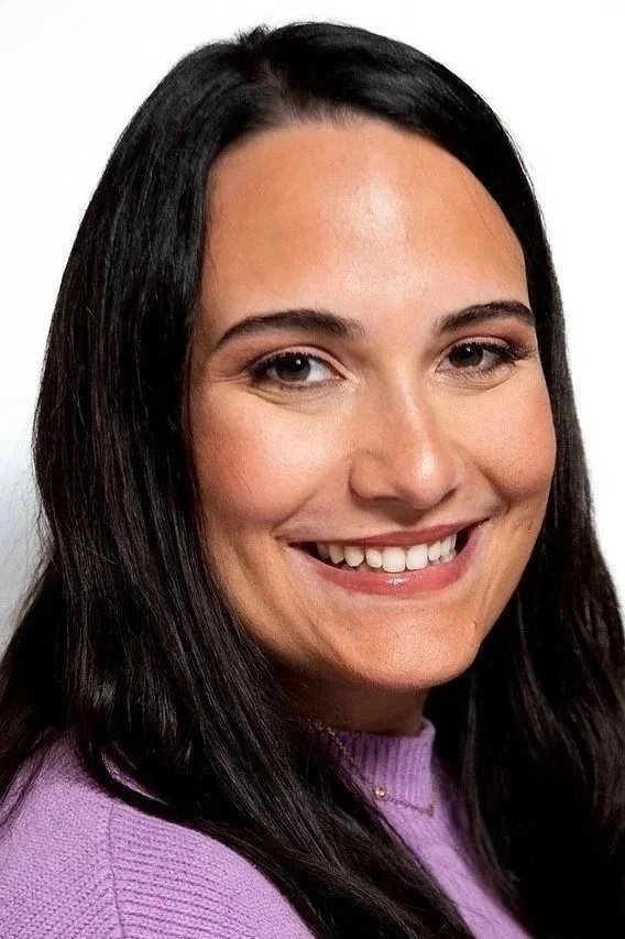 Close-up of a young woman with long dark hair wearing a purple sweater, smiling at the camera against a white background.