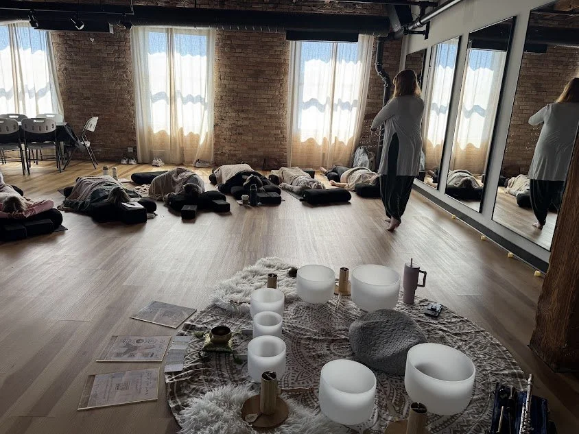 People lying on yoga mats in a spacious studio with large windows, participating in a sound healing session with crystal bowls and candles, led by an instructor.