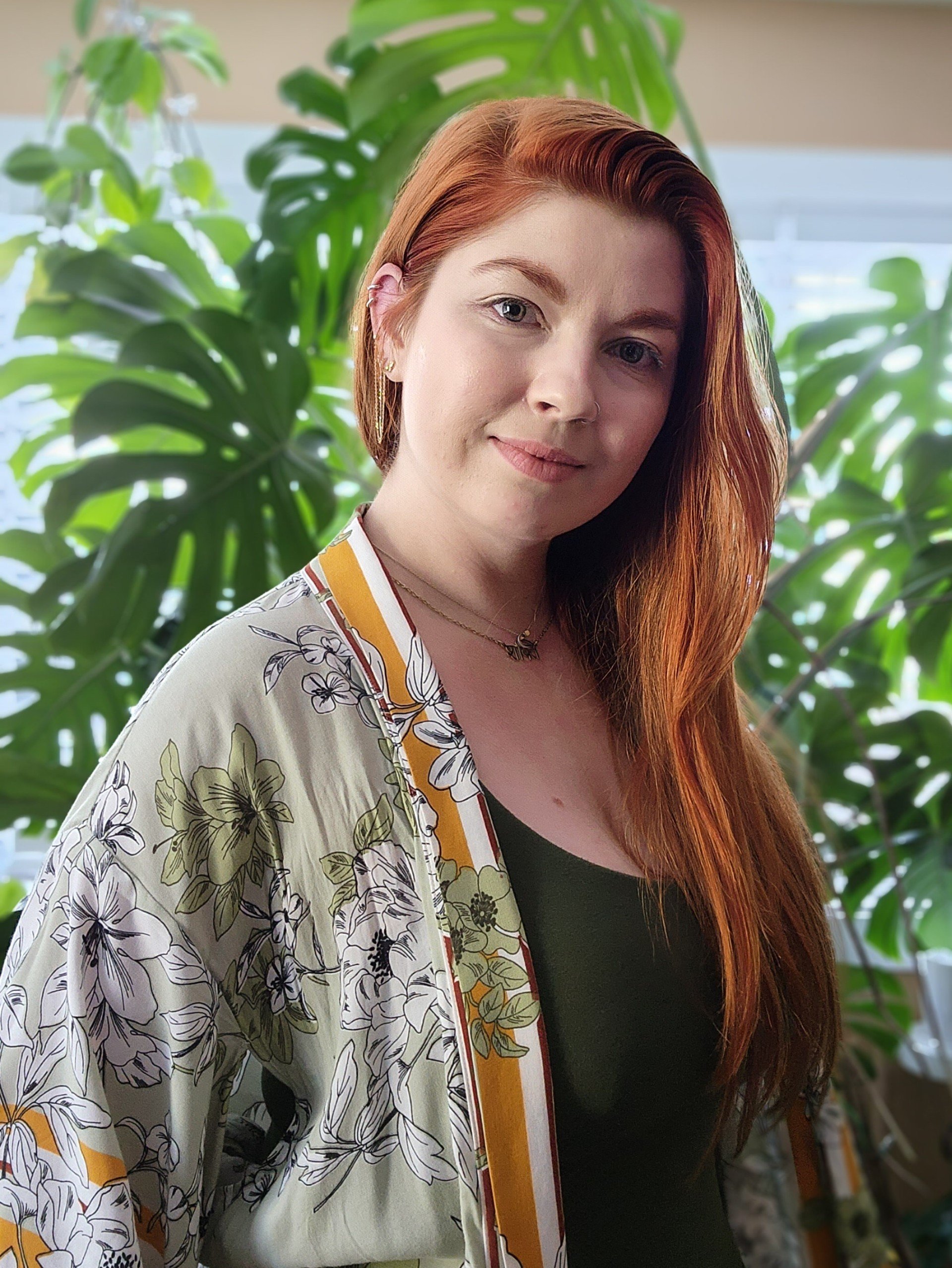 A woman with long red hair, wearing a floral patterned kimono-style jacket over a black top, standing in front of large green tropical leaves in an indoor setting with natural light.