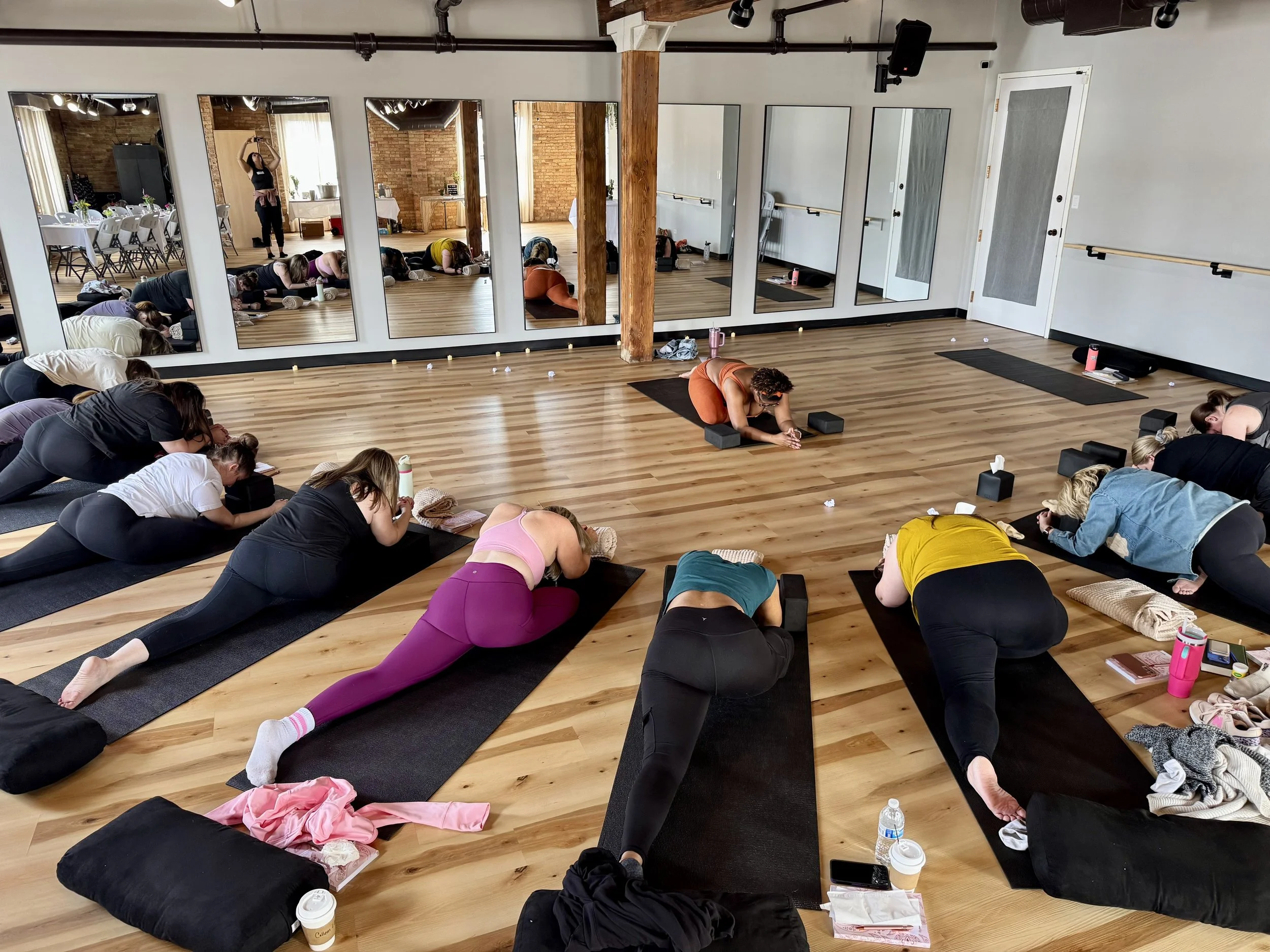Group of people participating in a yoga or meditation class in a spacious, wooden-floored studio with large mirrors on the wall.