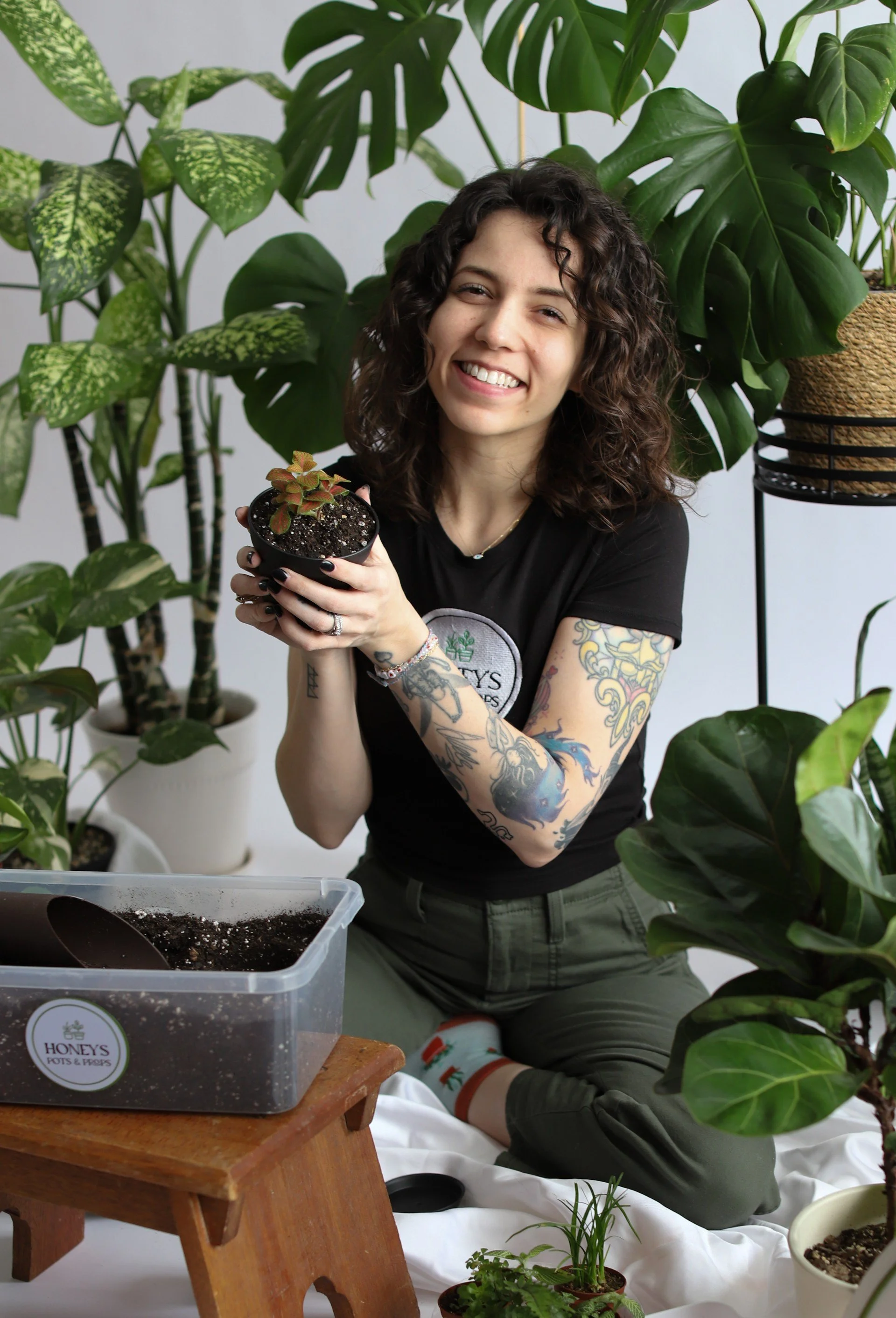 A woman with curly hair and tattoos smiling while holding a small potted plant in a room filled with various large green houseplants.