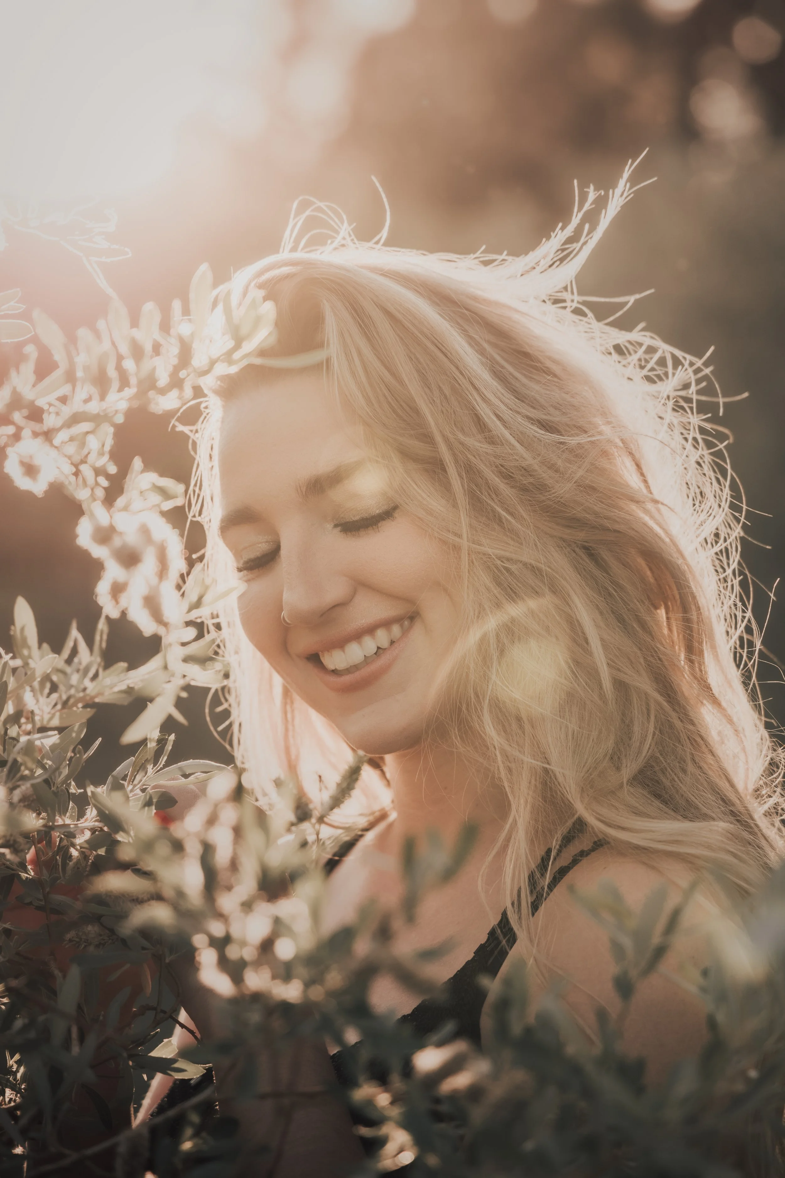A woman with blonde hair smiling with her eyes closed, surrounded by plants, with warm sunlight illuminating her face and hair.