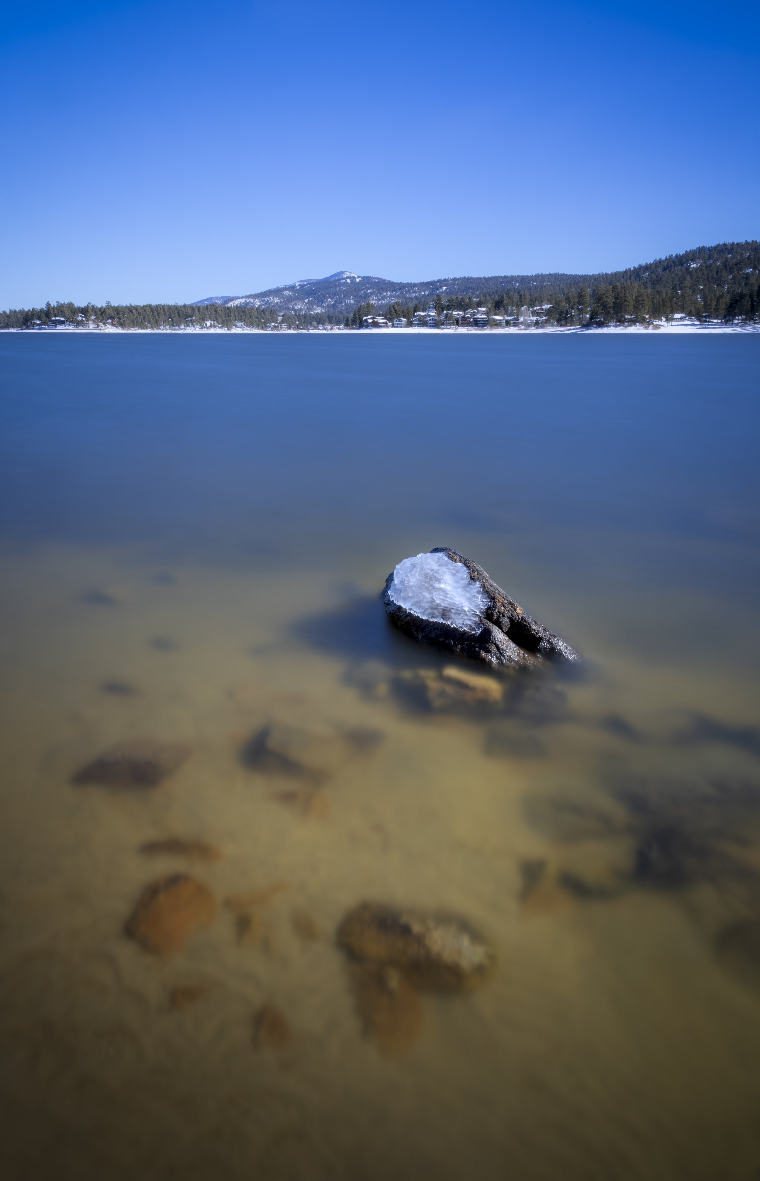 A snowy landscape featuring a lake with a partially submerged rock in the foreground, snow-capped trees along the shoreline, and mountains in the distance under a clear blue sky.