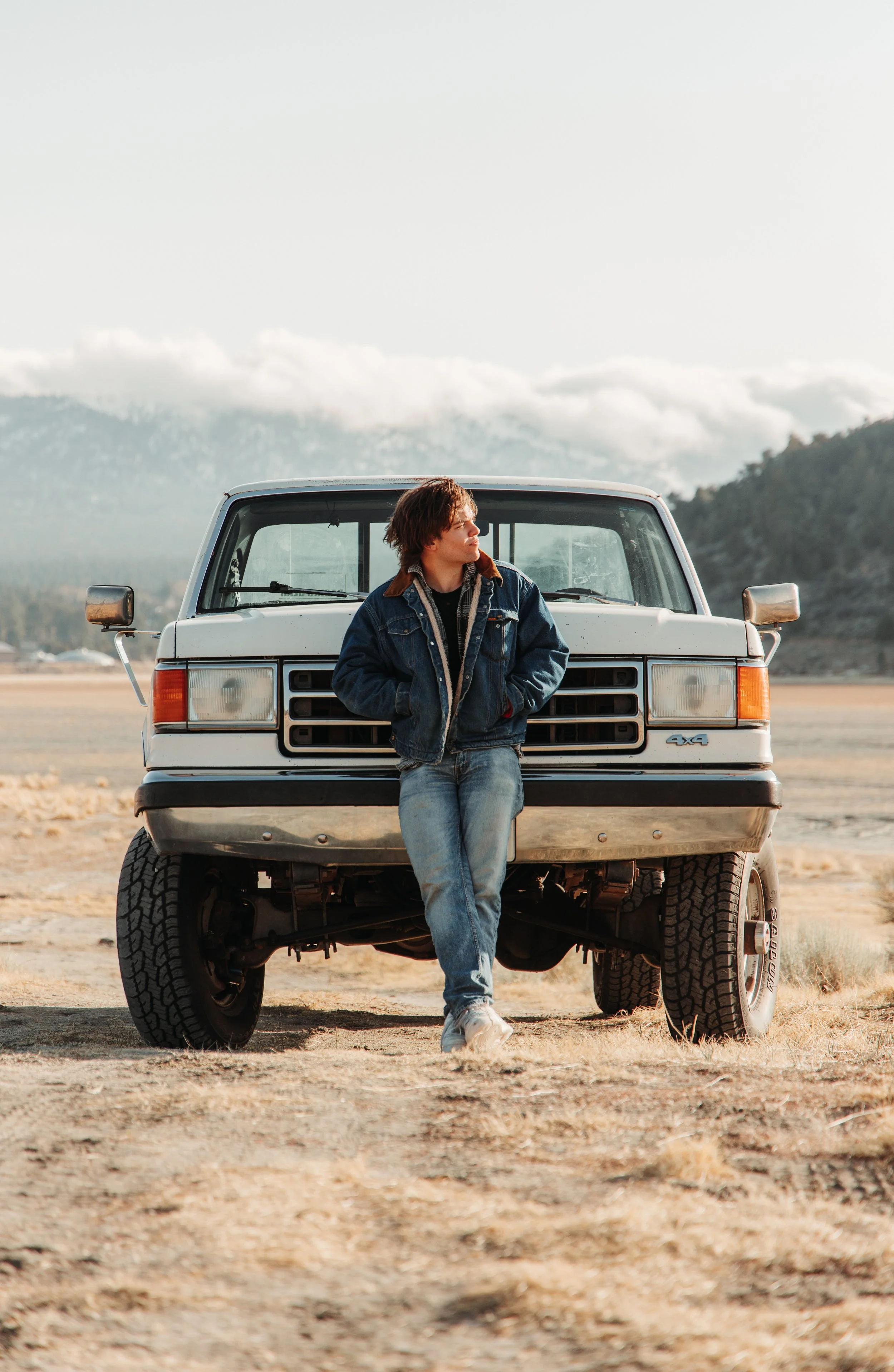 A young man in a denim jacket and jeans leaning against a white off-road vehicle in an open, dry landscape with mountains in the background.