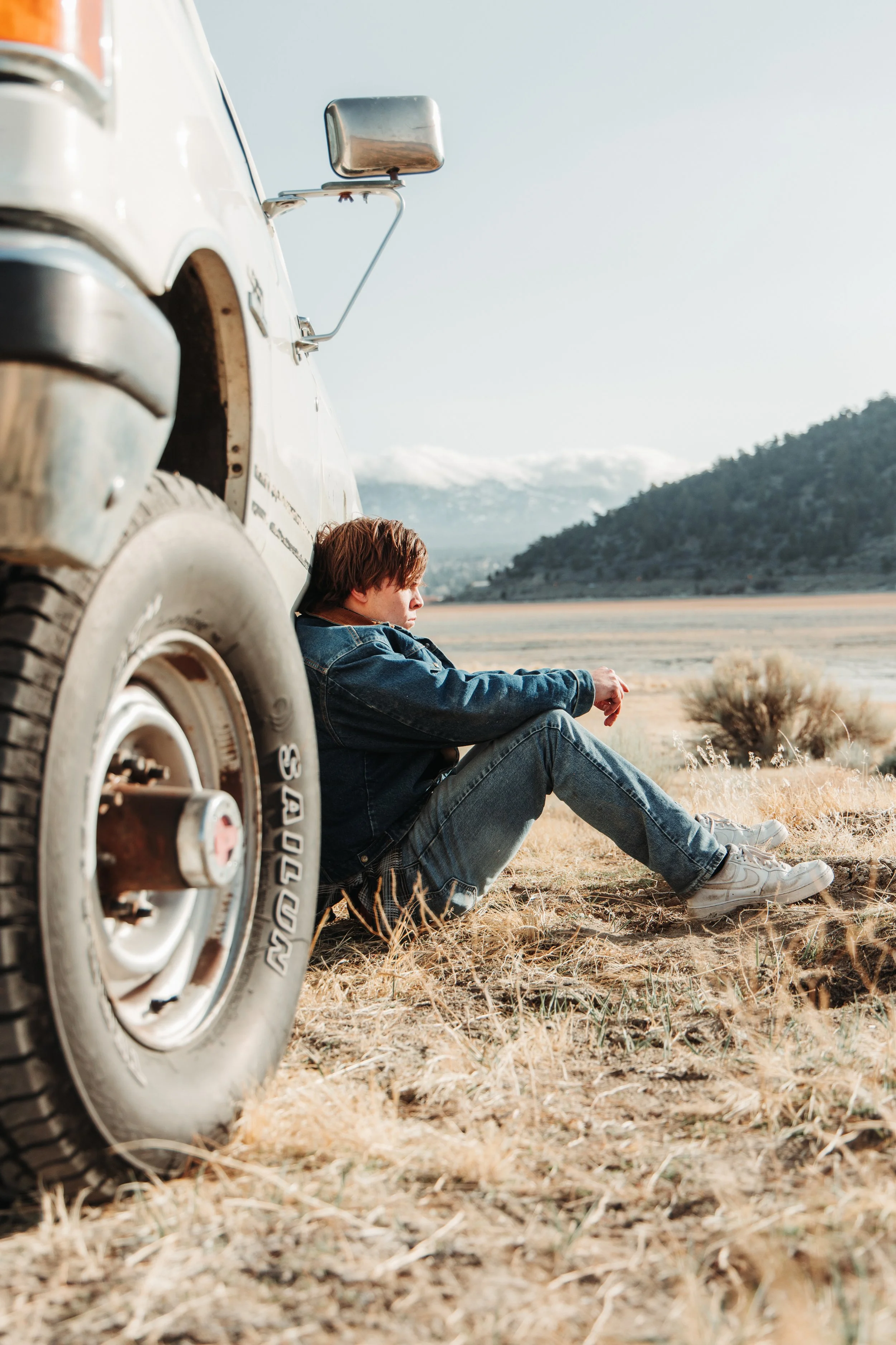 A person sitting on the ground leaning against a vintage off-road vehicle in a dry, open landscape with mountains in the background.