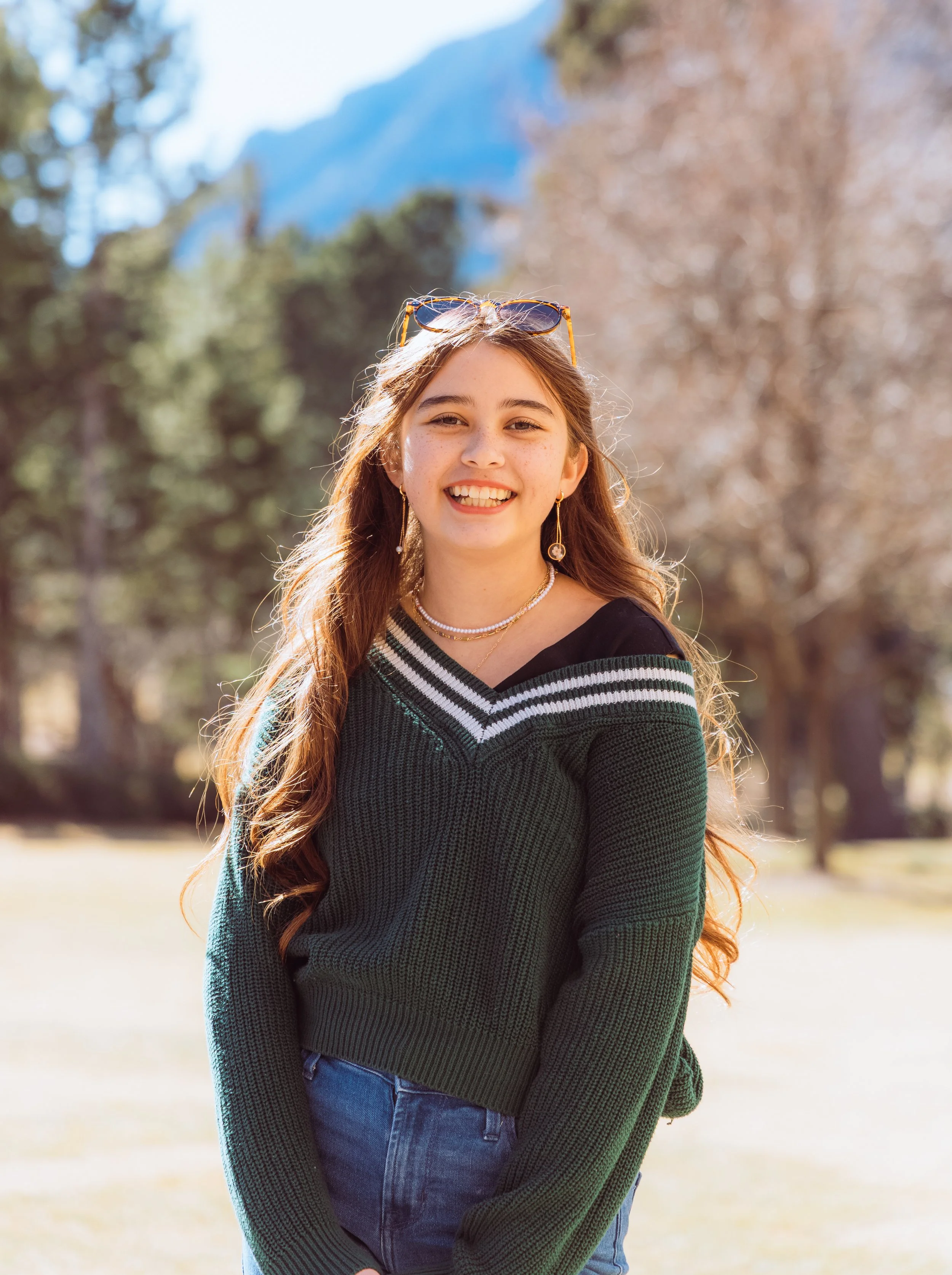 A young woman with long wavy hair smiling outdoors, wearing sunglasses on her head, a black and green striped sweater, blue jeans, and jewelry, with trees and mountains in the background.