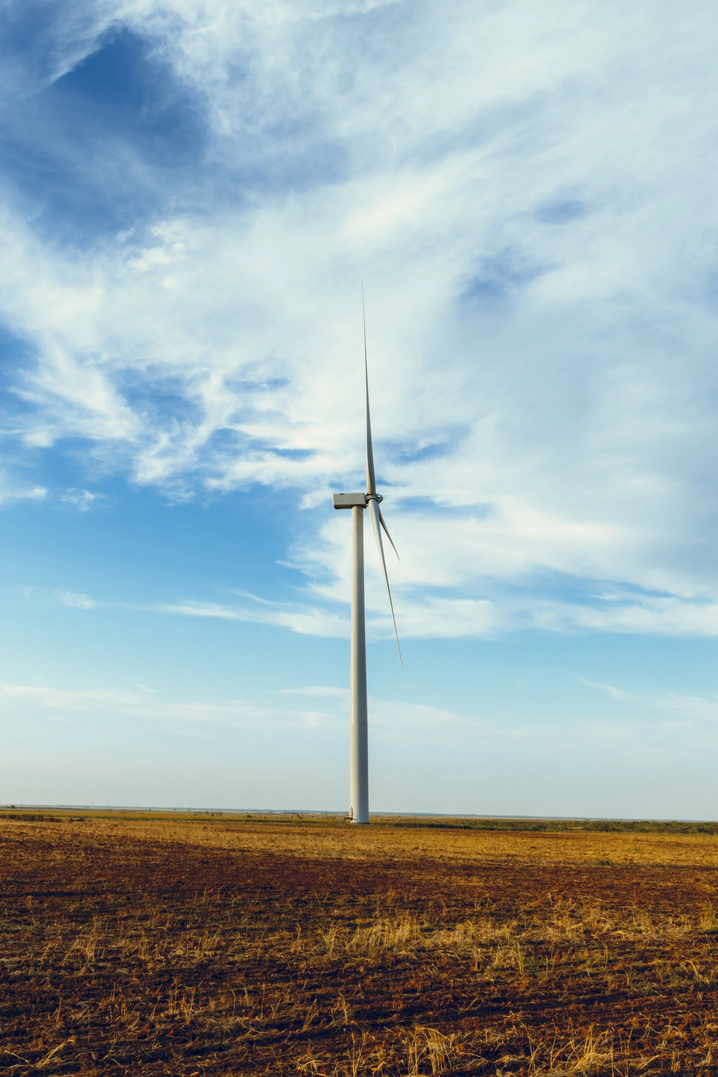 A single wind turbine stands in a dry, open field under a partly cloudy sky.