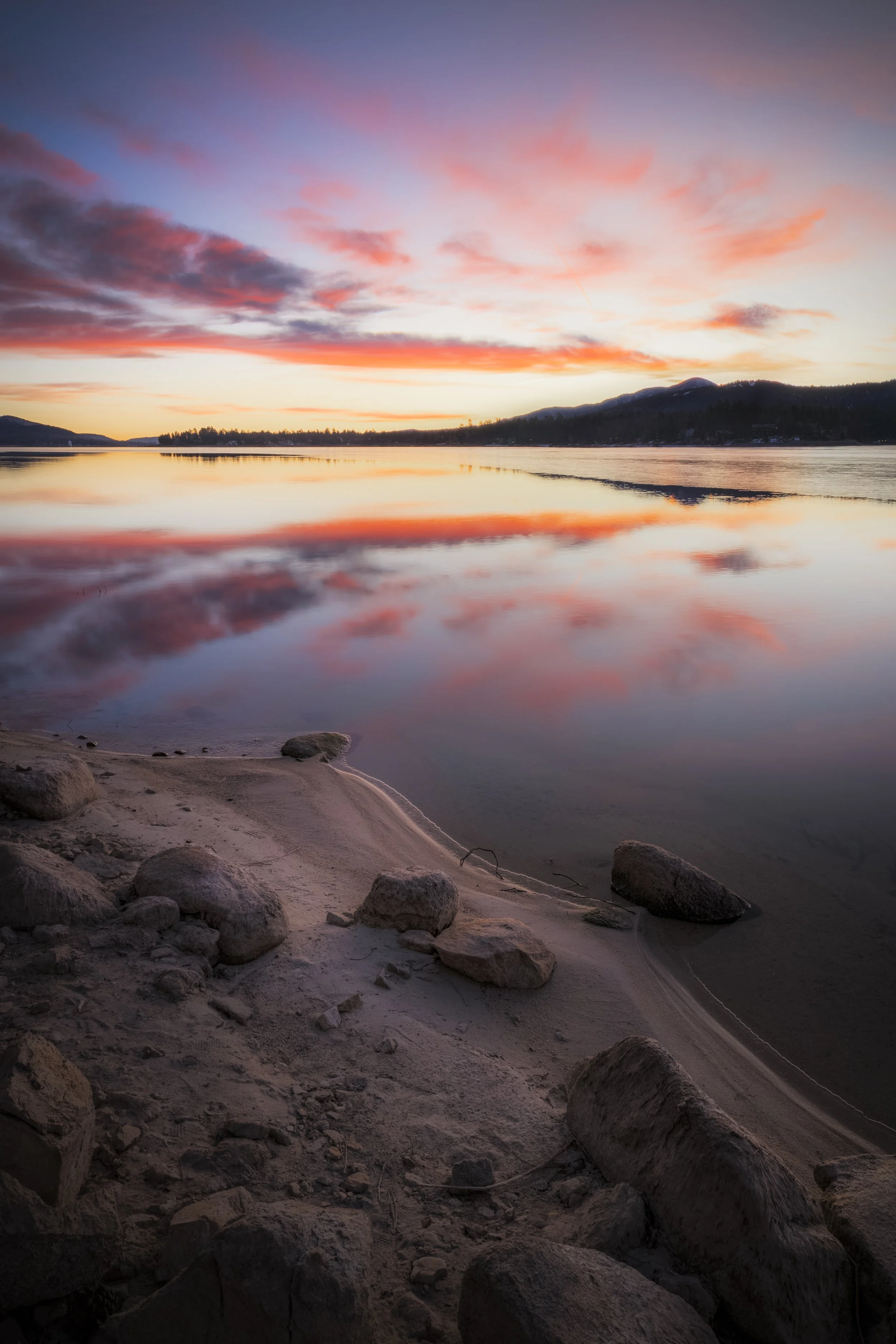 Sunset over a calm lake with pink and orange clouds reflected in the water, sandy shore with rocks in the foreground, mountain range in the background.