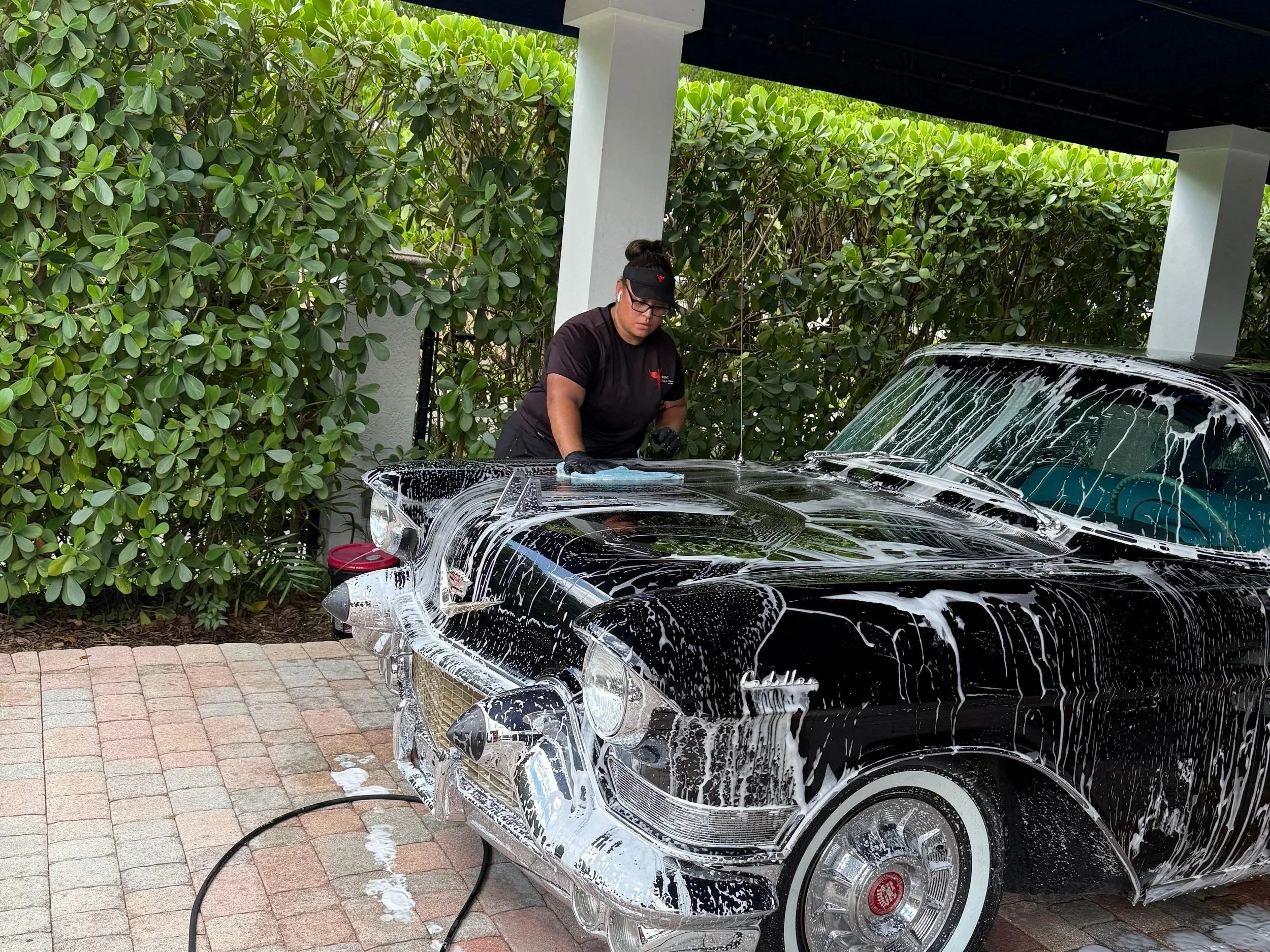 A person is washing a classic black Cadillac car, which is covered in white soap suds, under a carport with green bushes in the background.