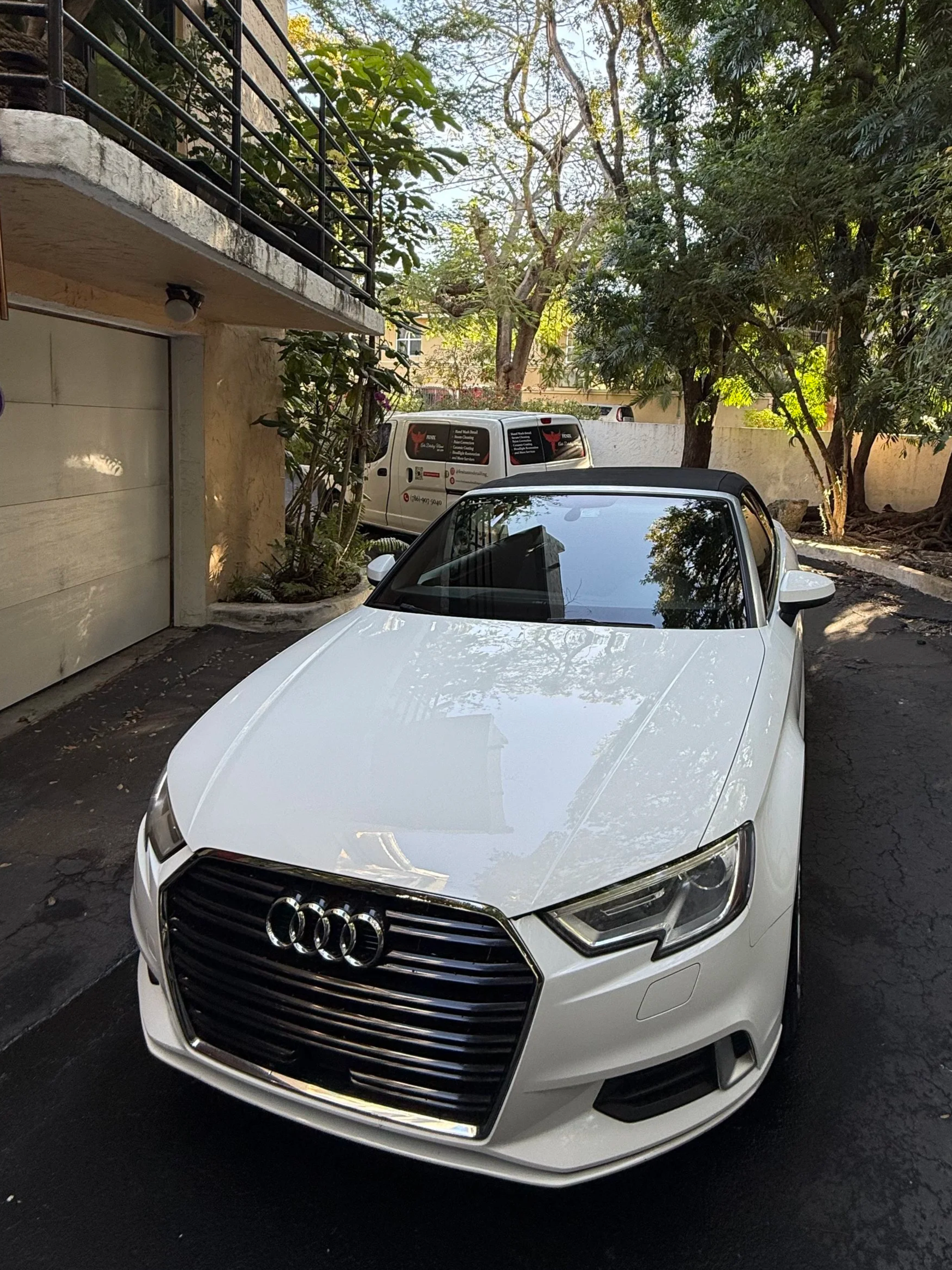 A white Audi convertible parked in a driveway surrounded by trees and a beige building with a garage door.