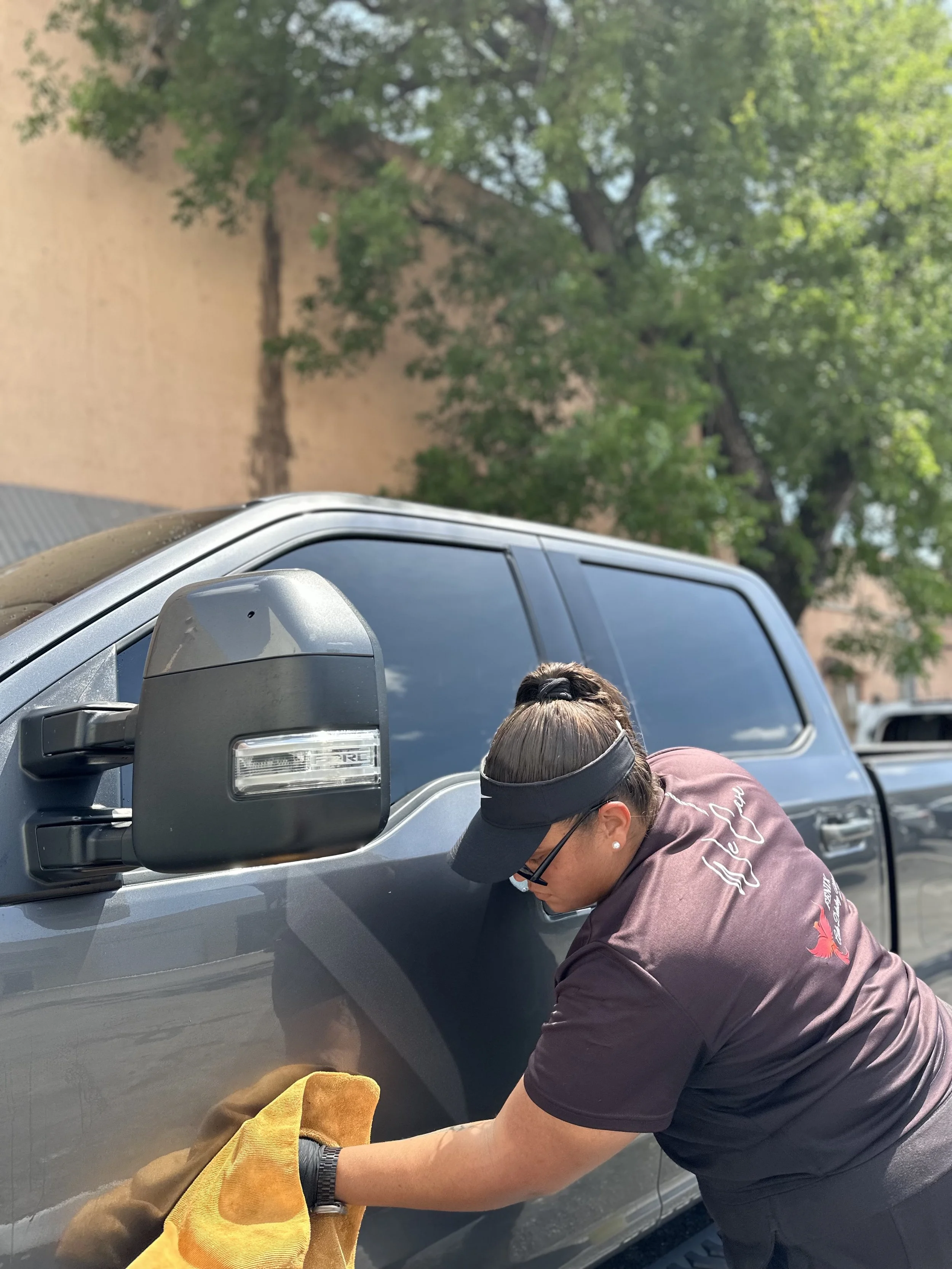 A woman cleaning a gray pickup truck with a yellow cloth. She is wearing a black visor, sunglasses, and a dark-colored shirt, standing outdoors with trees and a wall in the background.