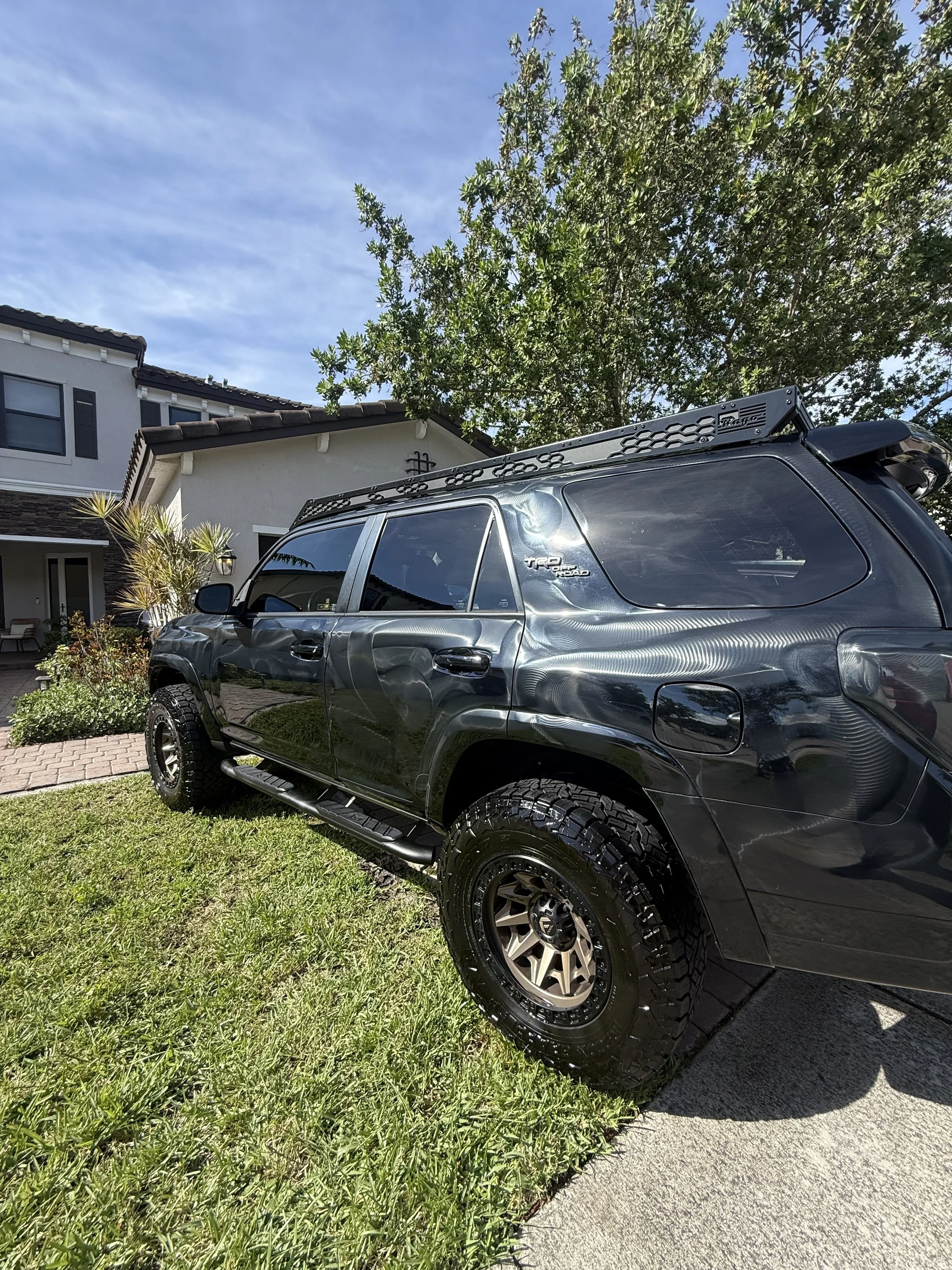 Black SUV parked on the grass next to a sidewalk in front of a house with a white facade, brown roof, and plants, under a clear blue sky.
