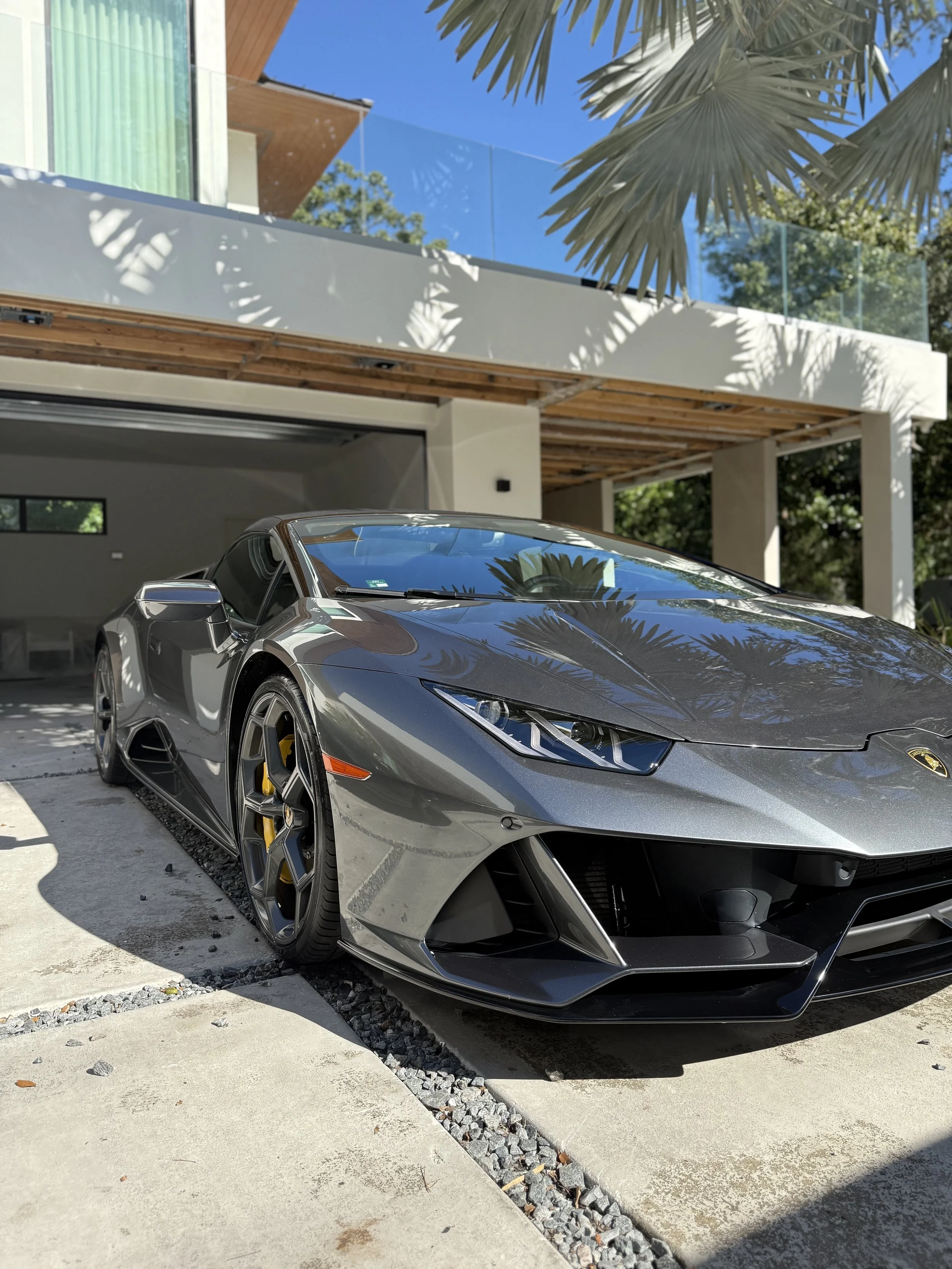 A sleek gray Lamborghini sports car parked in front of a modern house with a garage, glass balcony, and palm trees.