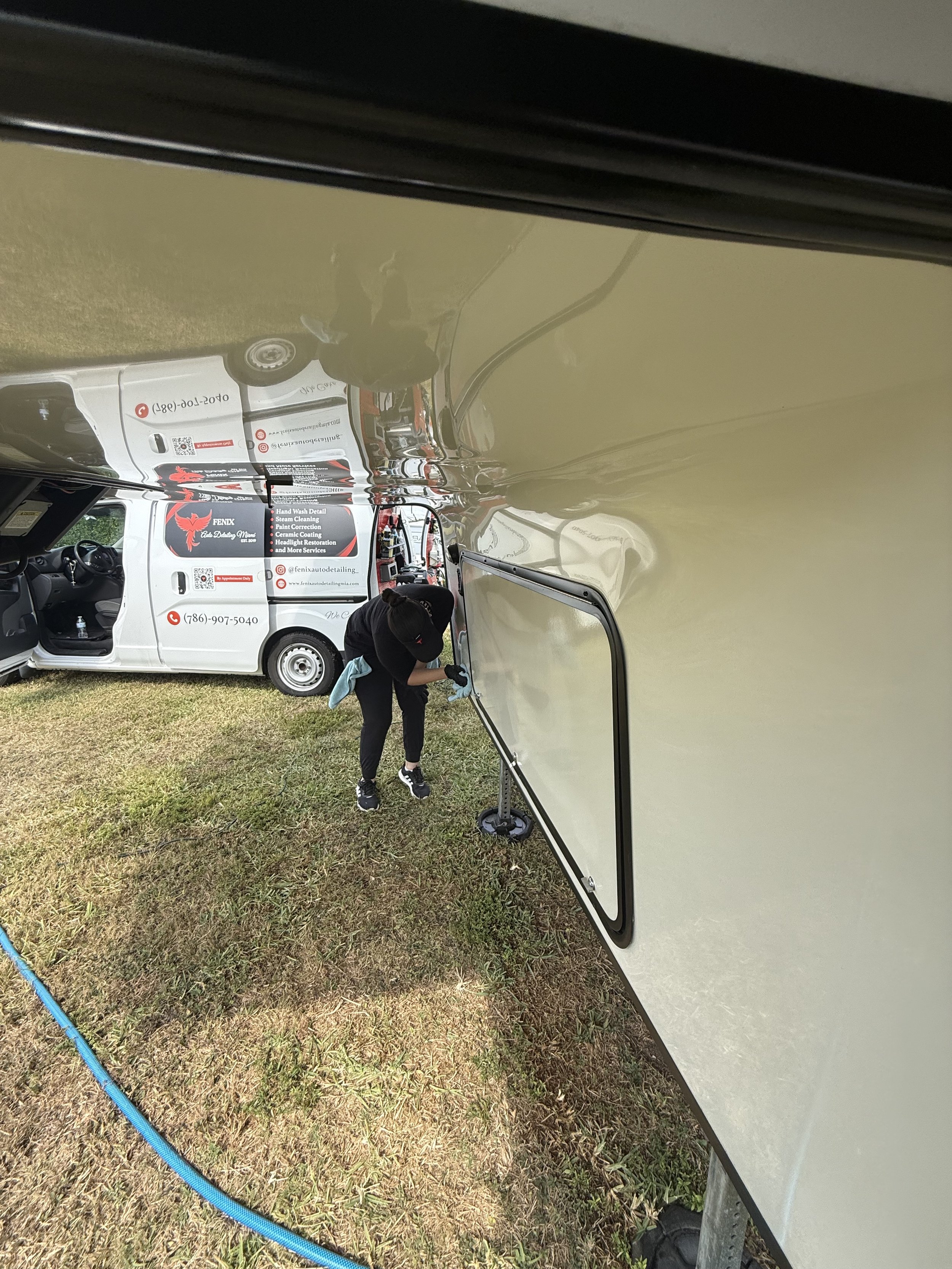 Person cleaning or inspecting a side window of a large RV or camper trailer, with a white van parked nearby that has signs and contact information for a detailing business.