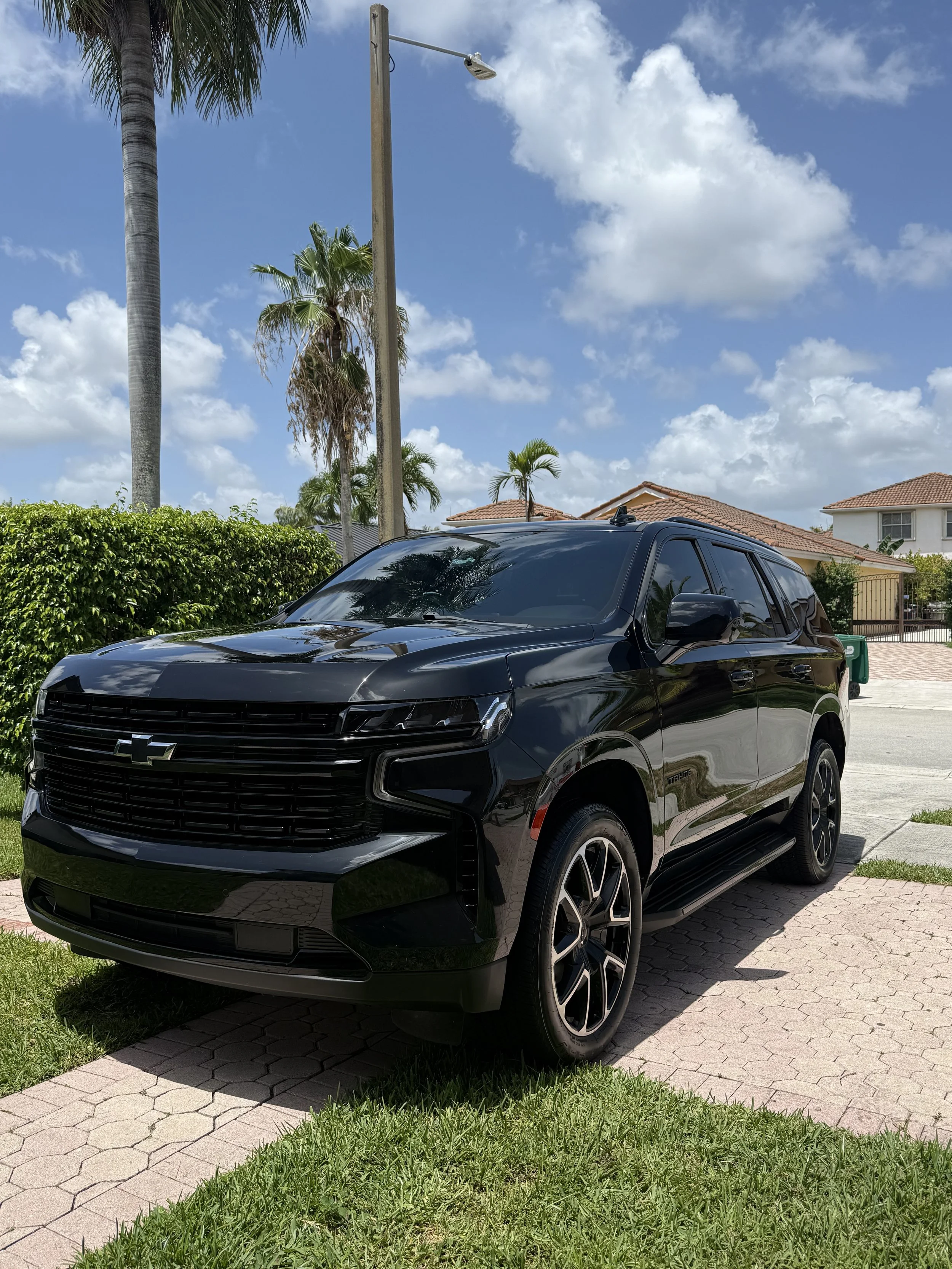 A black Chevrolet SUV parked on a driveway with a green lawn, palm trees, a hedge, and houses in the background under a partly cloudy sky.