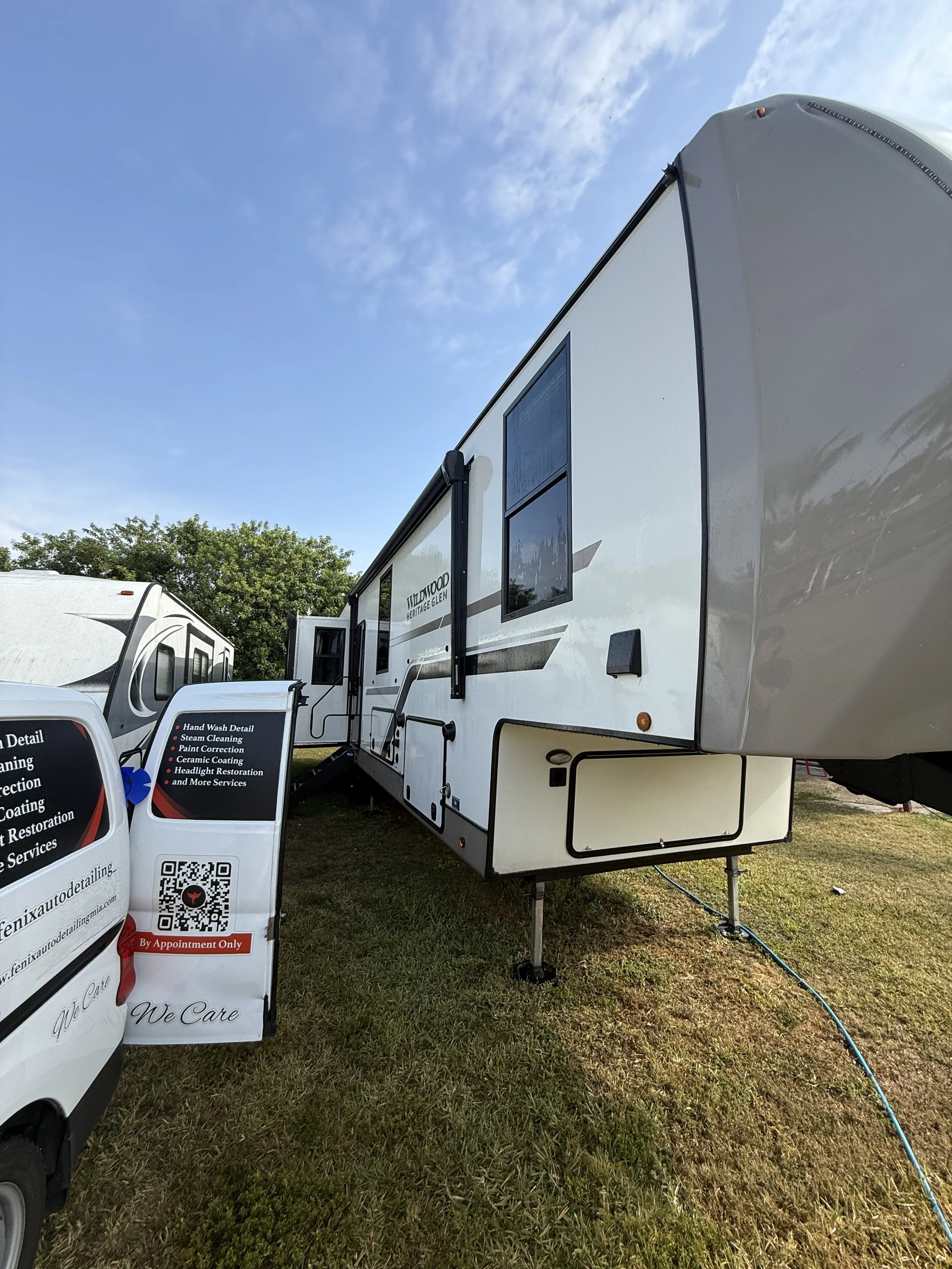 A white travel trailer parked on grass with a small signboard beside it, against a background of trees and a partly cloudy blue sky.