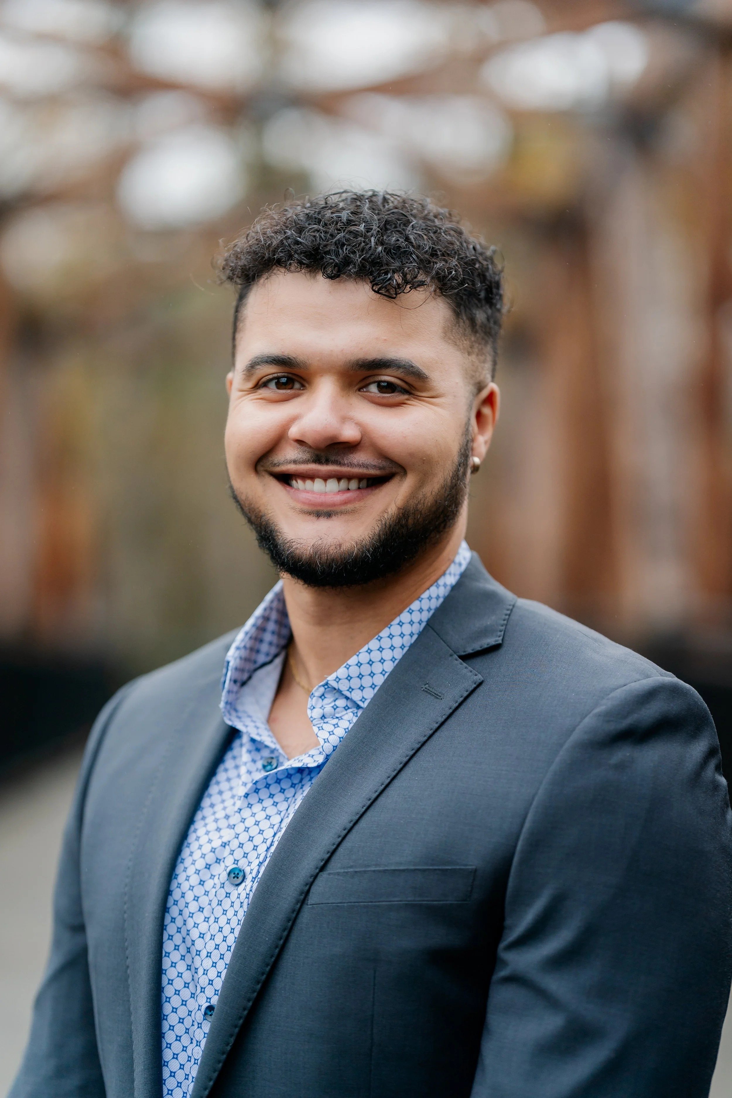 Smiling young professional man wearing blazer poses outdoors, showcasing confident personality through polished business headshot with natural background.