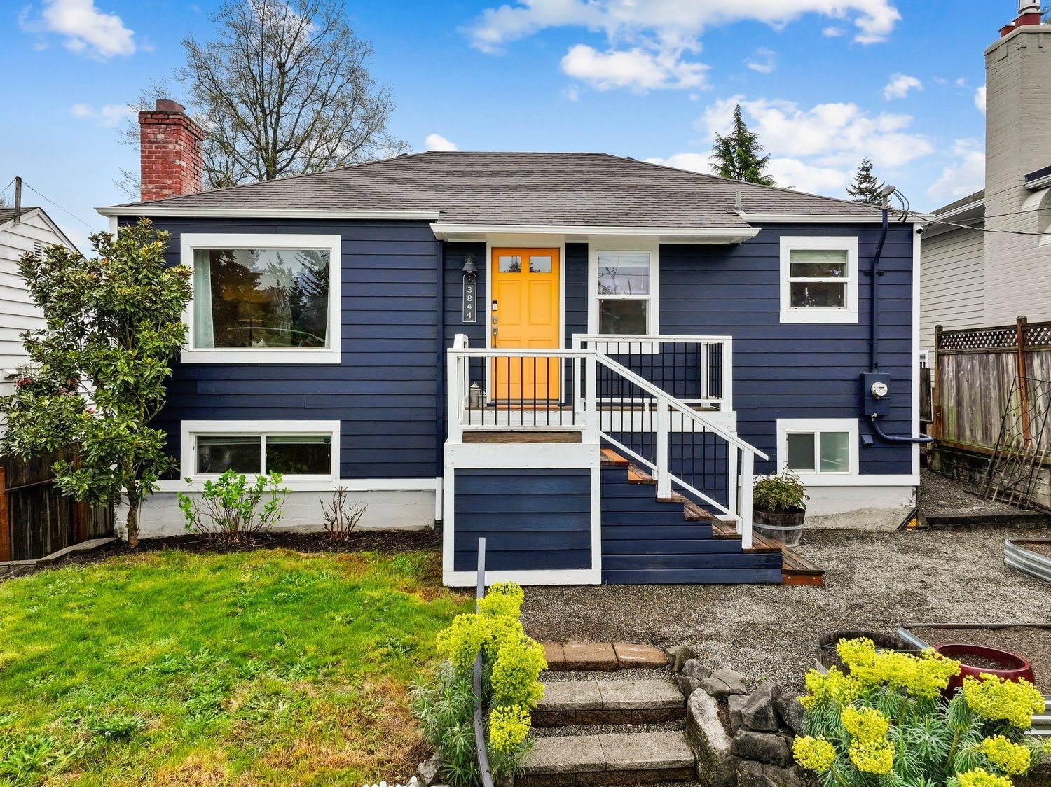 Charming blue cottage with bright yellow door, white trim, landscaped garden, front steps, and sunny sky.