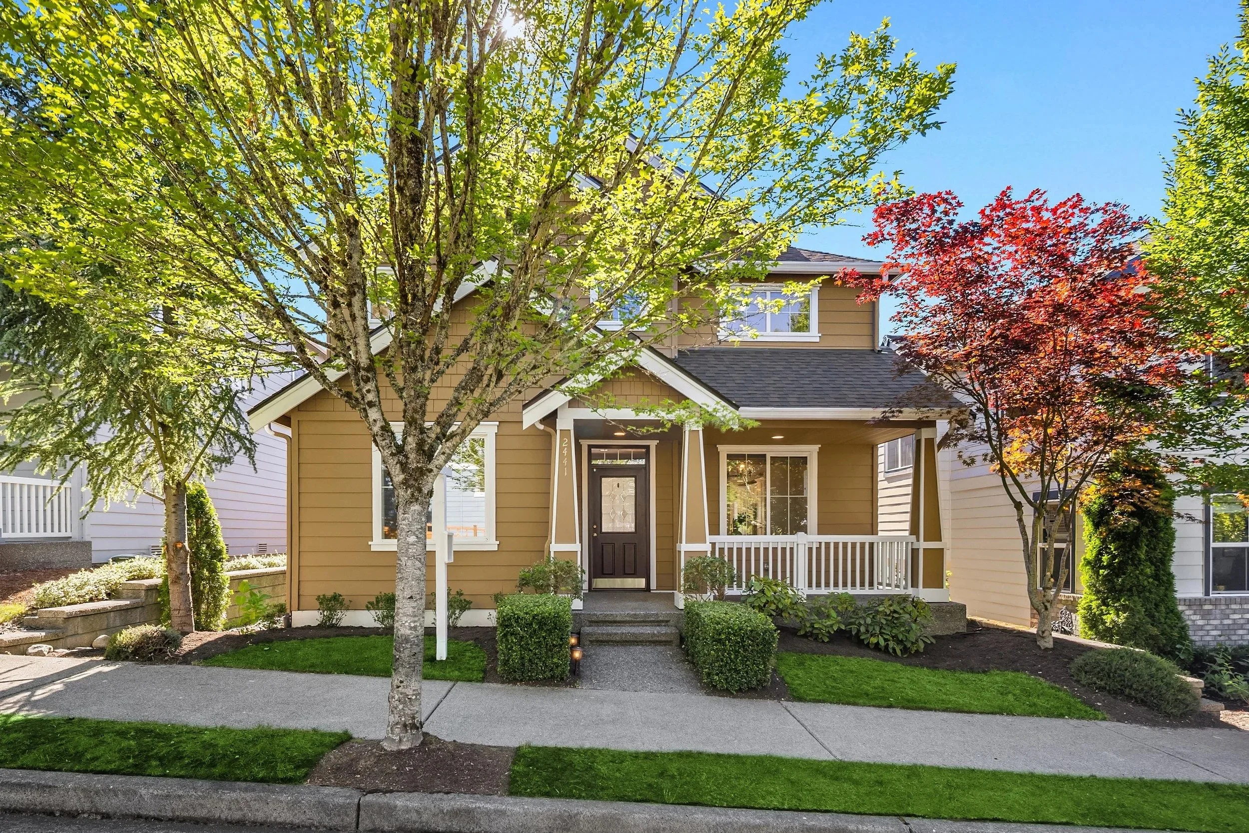 Charming suburban home with a welcoming porch, landscaped garden, mature trees, and bright sunny blue sky.