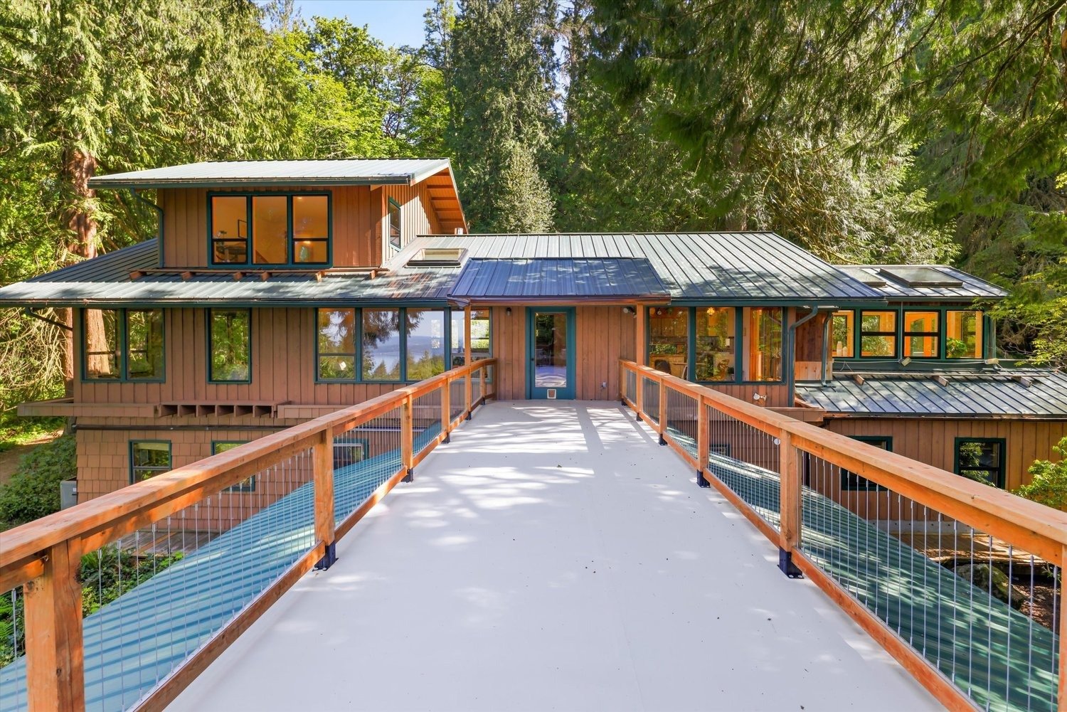 Modern woodland retreat with wood siding, expansive windows, metal roof, and elevated walkway surrounded by trees.