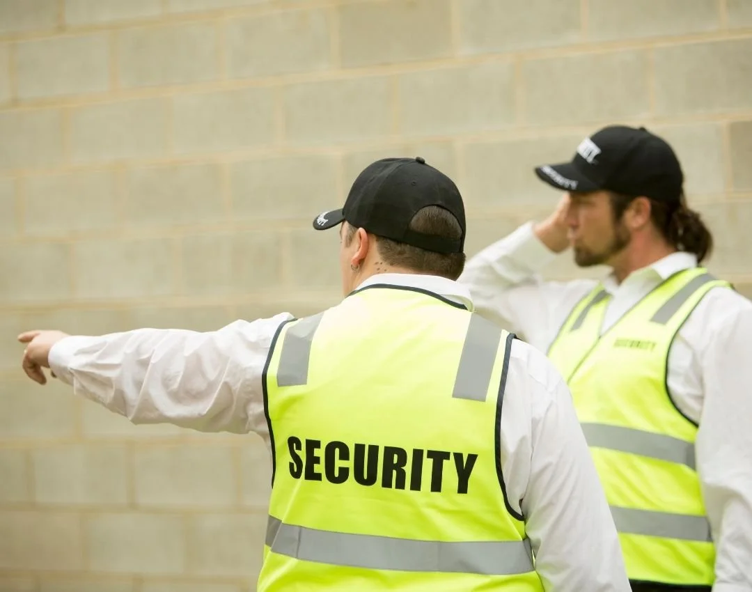 Two security guards in high-visibility vests and black caps, standing against a beige brick wall, with one pointing and the other adjusting his hat.