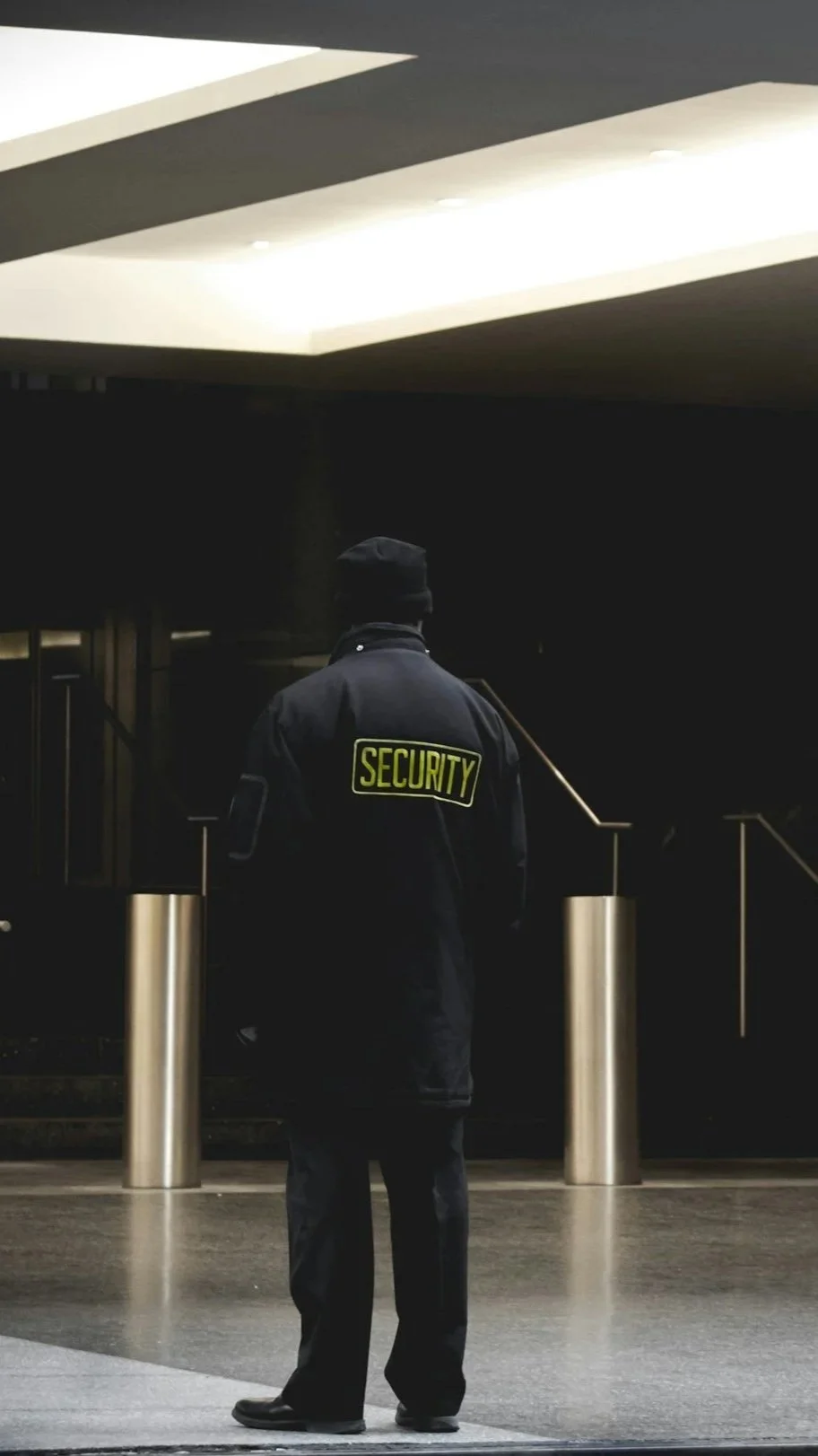 A security guard standing in a dark, indoor public space, facing away, wearing a black jacket with a yellow 'SECURITY' patch on the back and a black beanie, with modern architecture and metallic poles in the background.