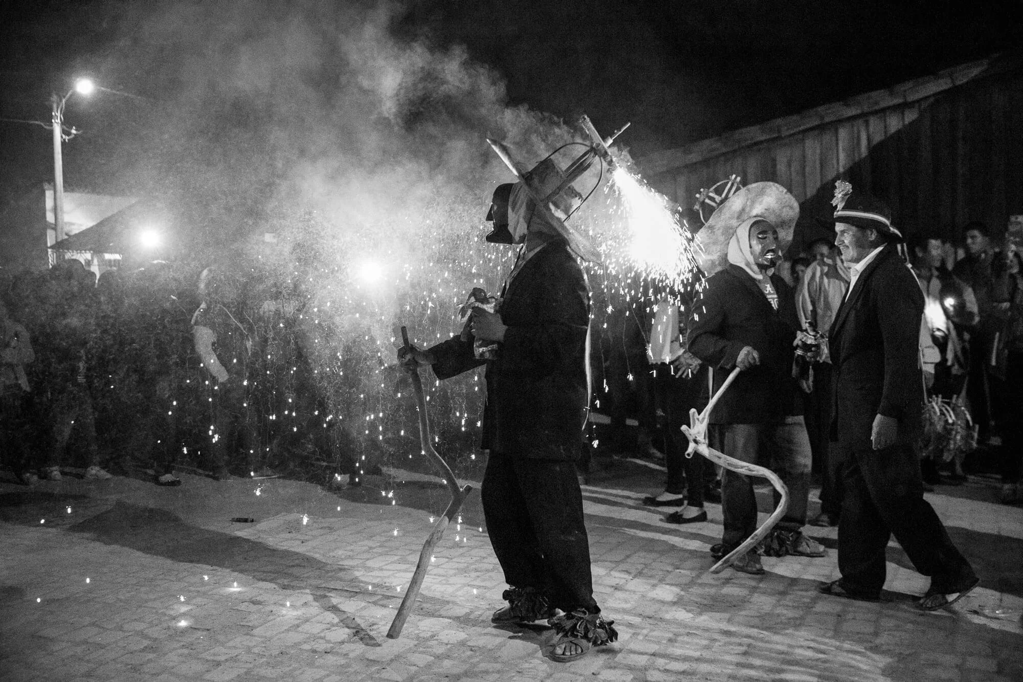 black and white photo of a festive crowd at night with two masked men in a central clearing lit by sparkling pyrotechnics