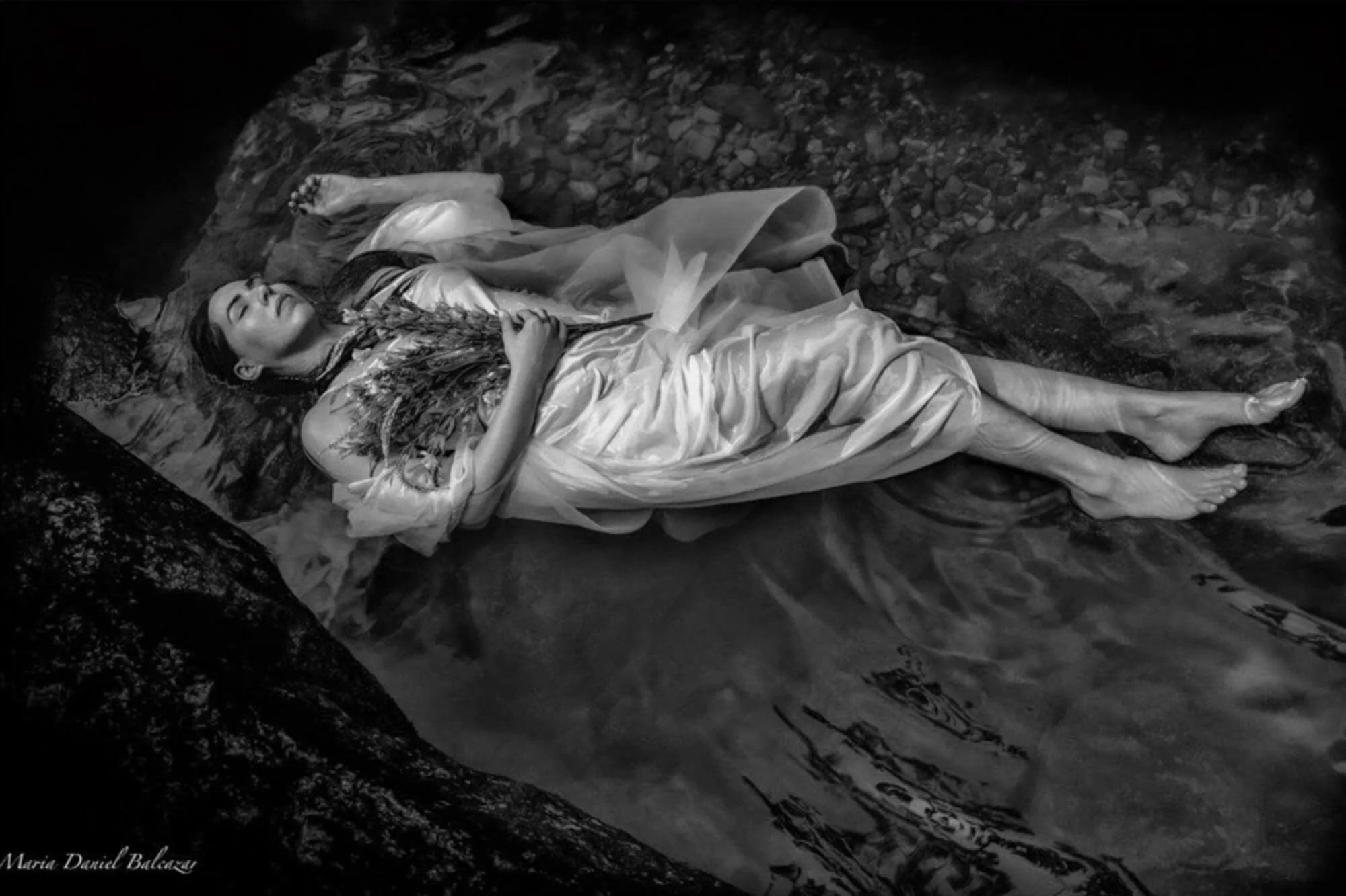 black and white photo of a women laying in a stream bed with her eyes closed wrapped in a translucent dress and holding a bouquet