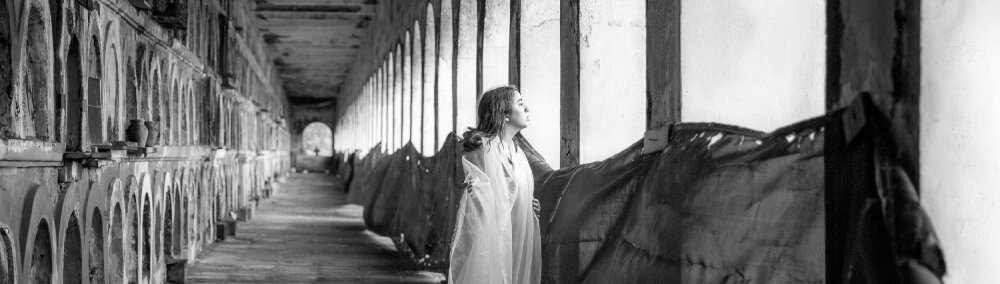 black and white photograph of a woman in a white dress staring into the light from inside an arch adorned hallway