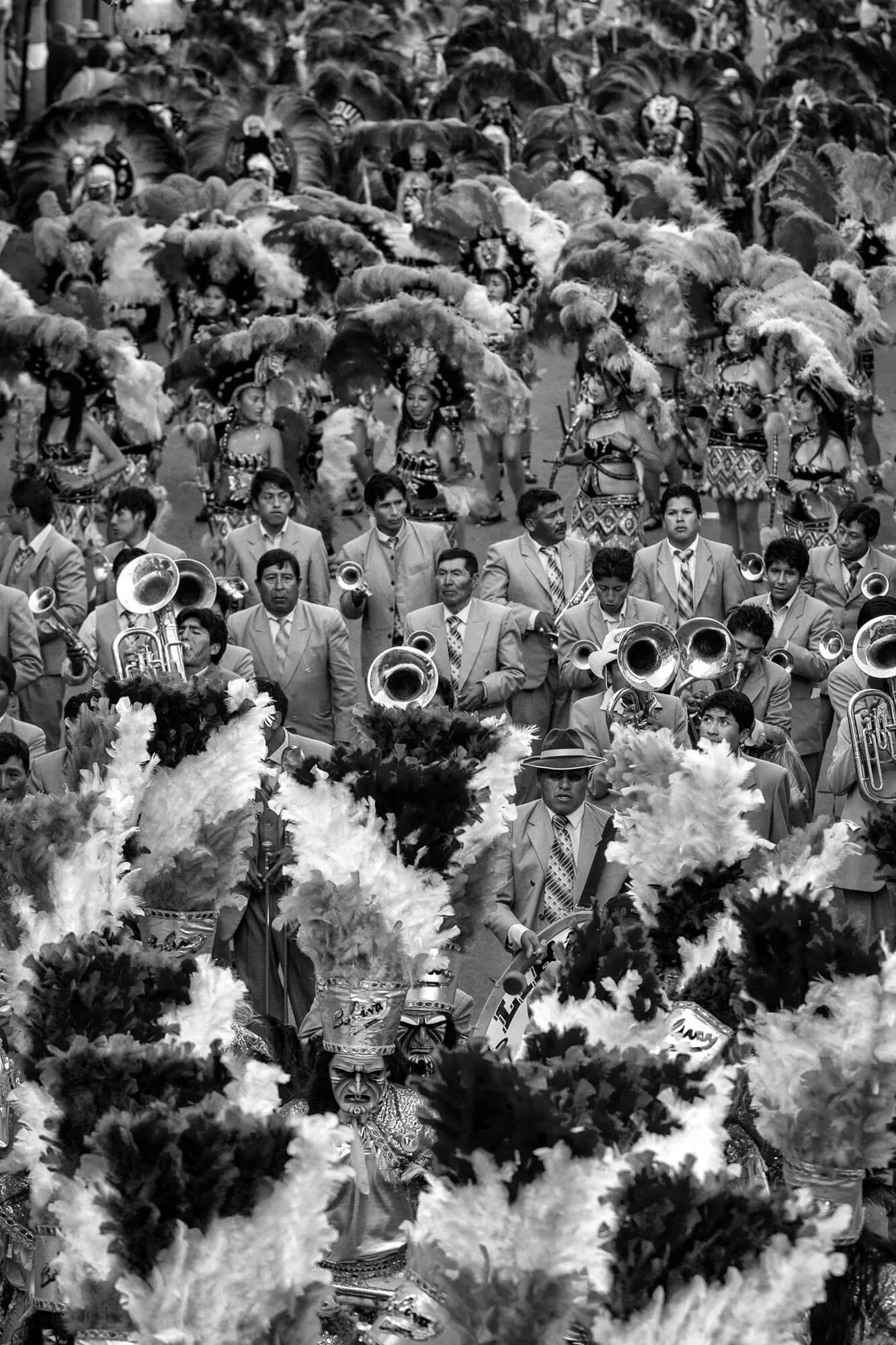 long focal length overhead black and white photo of an Oruro Carnival street parade showing layers of performers alternating between costumes with masks and large feathers and brass performers