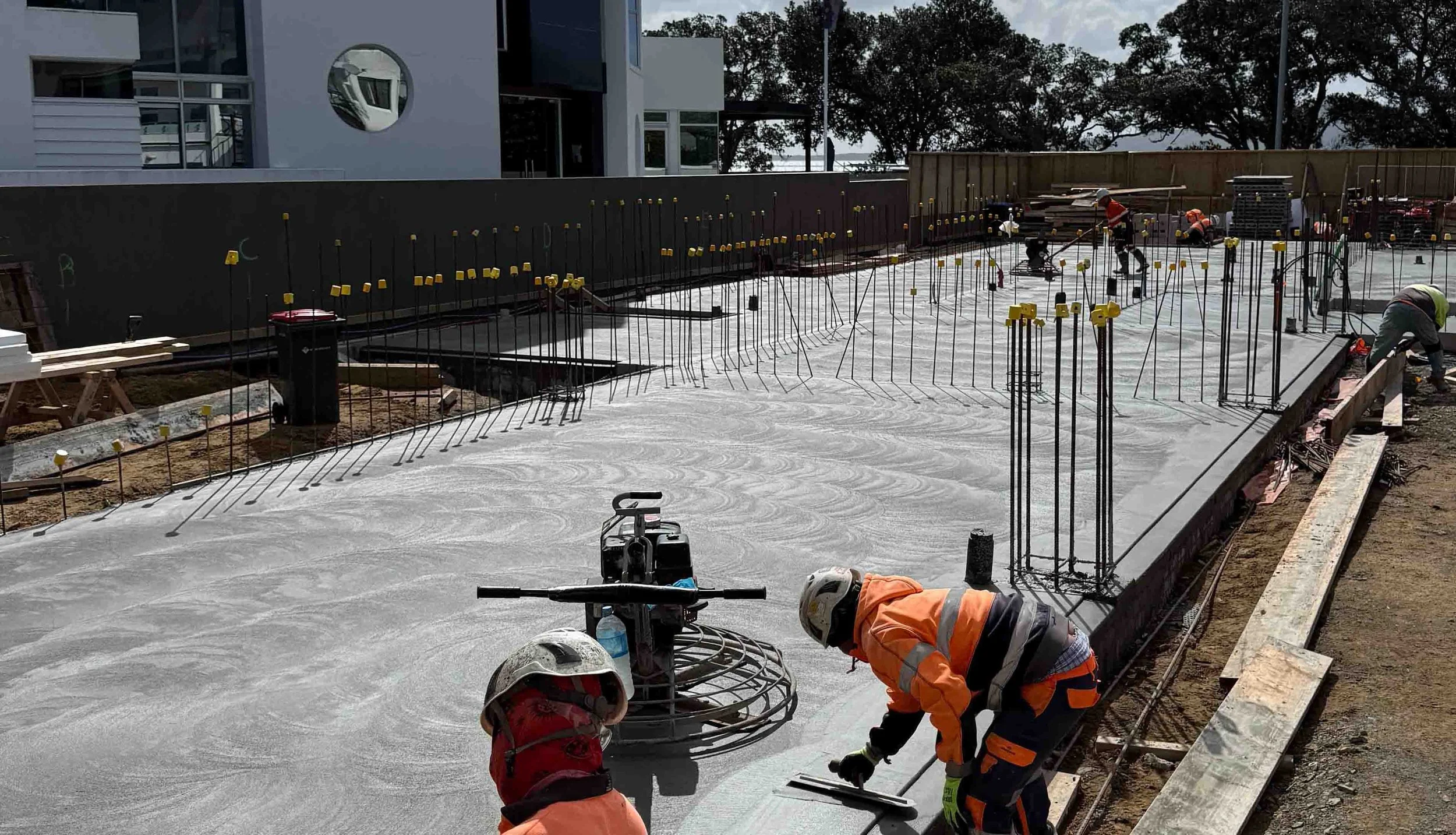 Construction site with workers pouring concrete, constructing foundation, and building a structure, surrounded by trees and with construction equipment and materials.