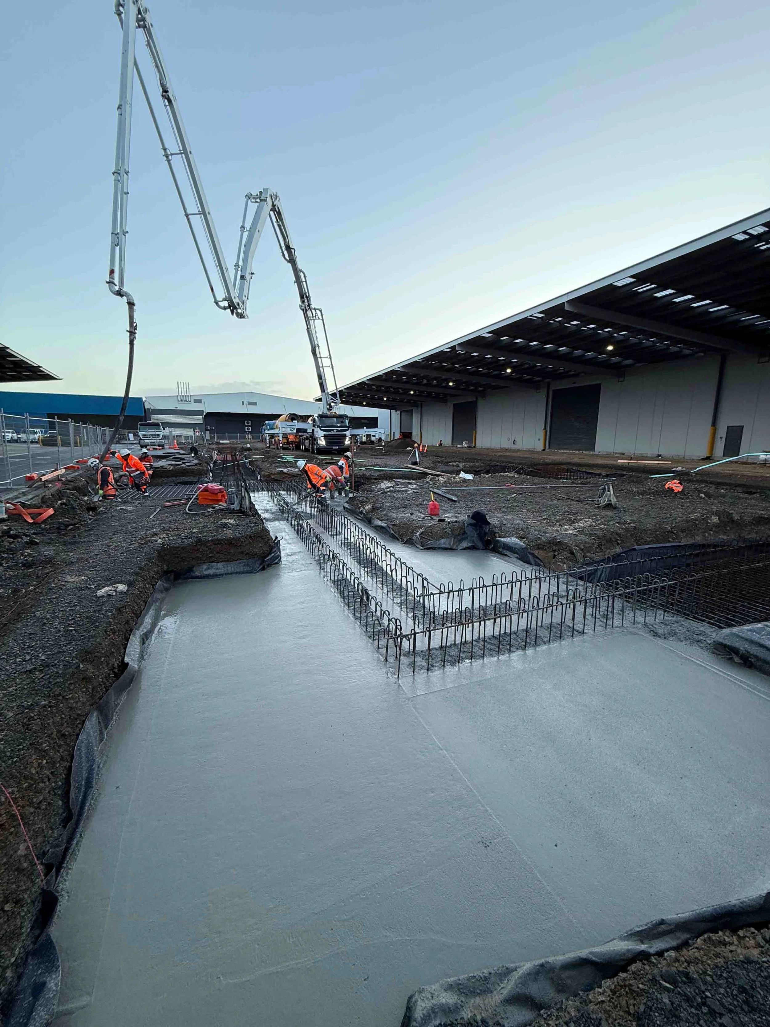 Construction workers pouring concrete on a construction site near large industrial buildings with a clear sky overhead.