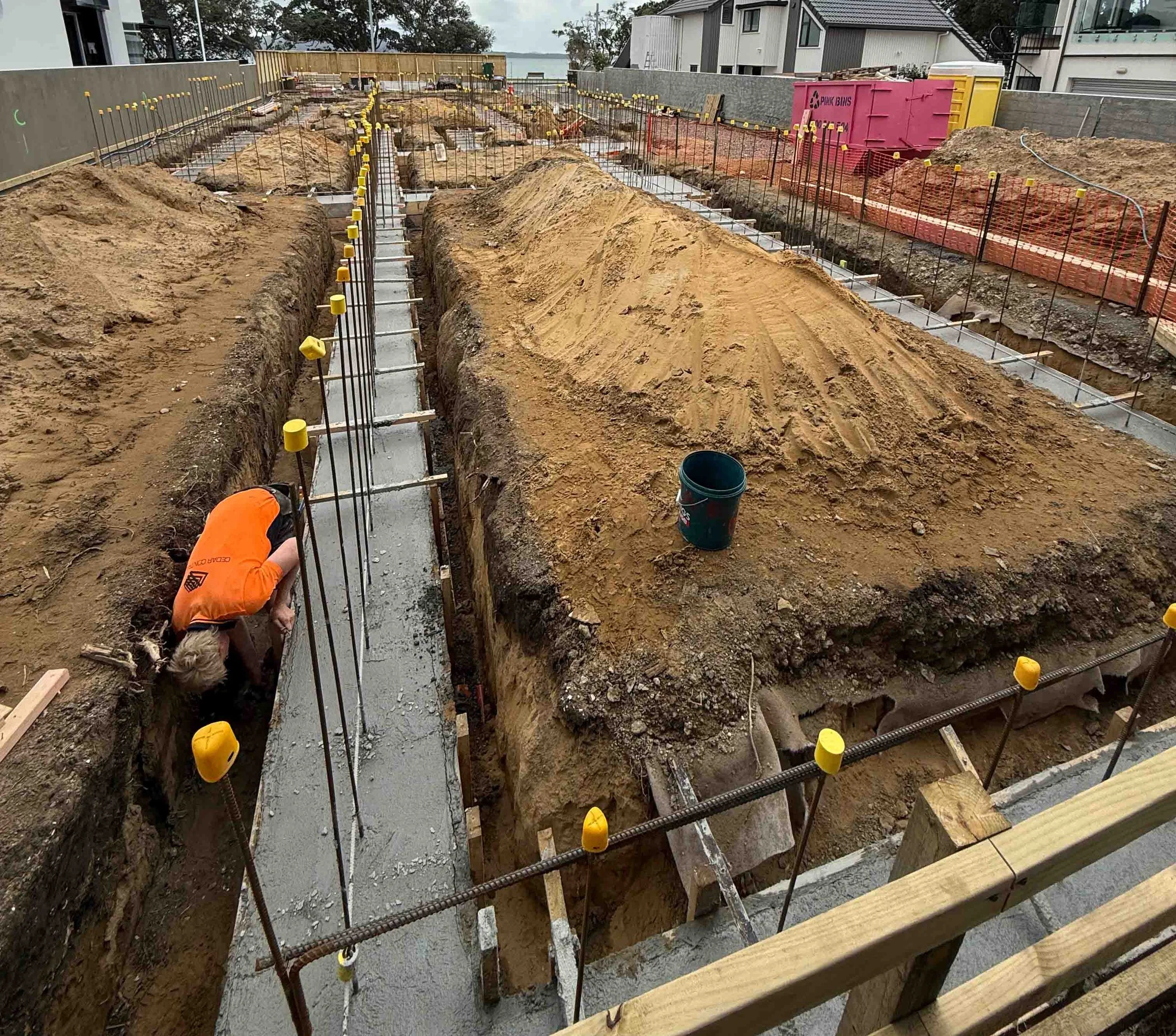 Construction site with trenches being prepared for foundational work, with a worker crouched down working on the concrete foundation, surrounded by dirt and construction materials.