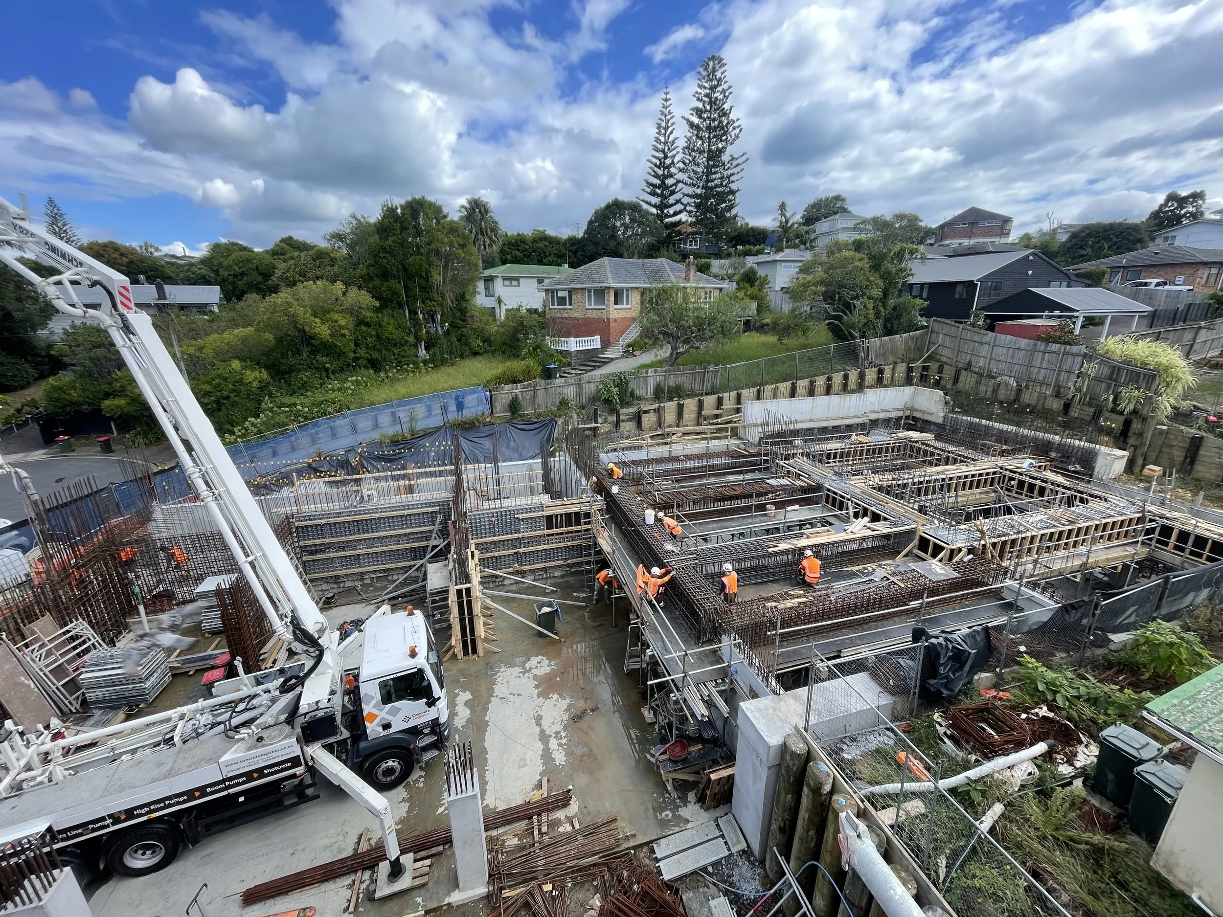 Construction site with workers in orange safety vests and helmets, concrete foundation being poured, and residential neighborhood in the background under a partly cloudy sky.