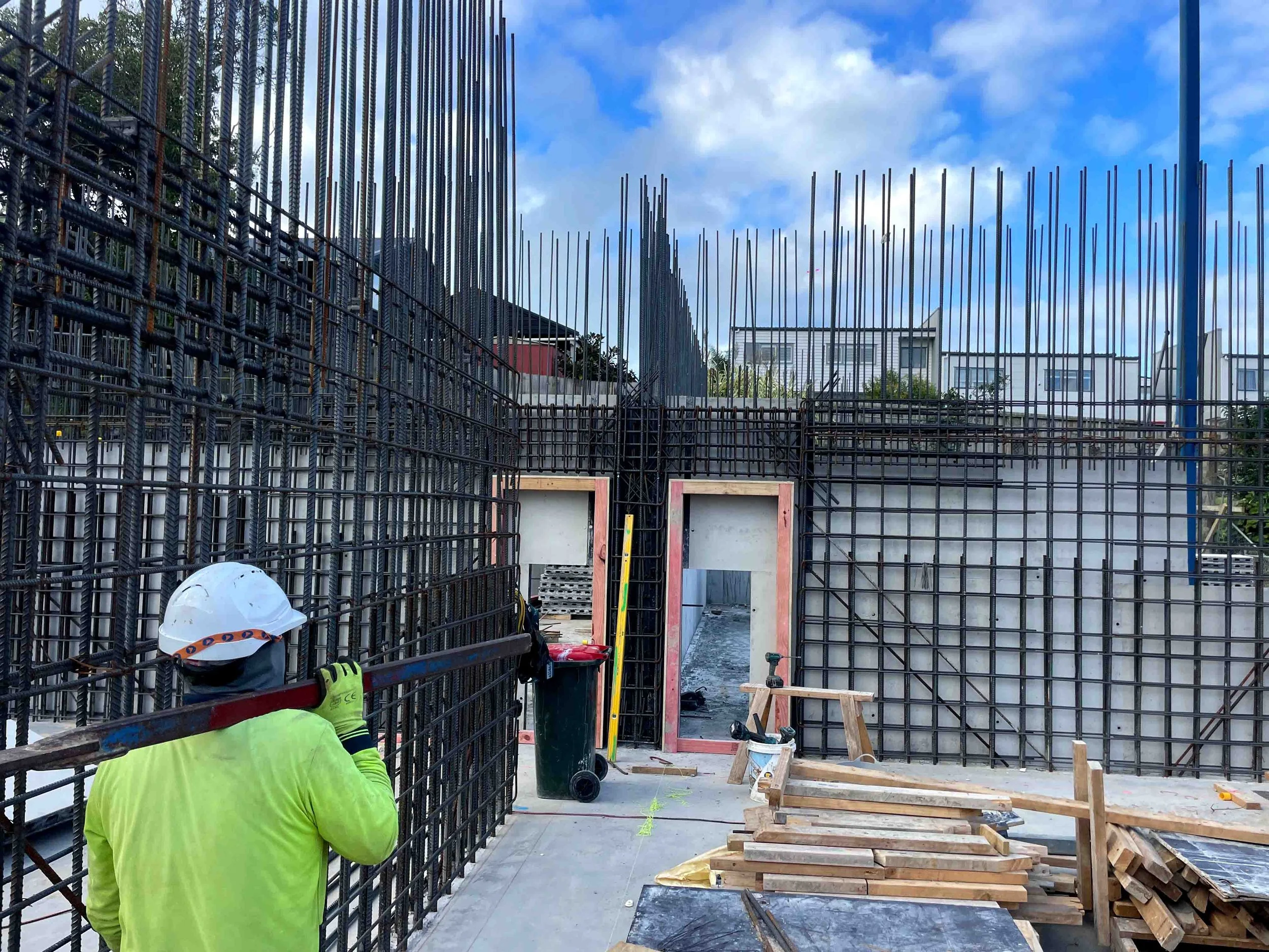Construction worker in neon yellow shirt and white helmet inspecting reinforced steel rebar framework at a building site. Construction tools and materials are scattered around.