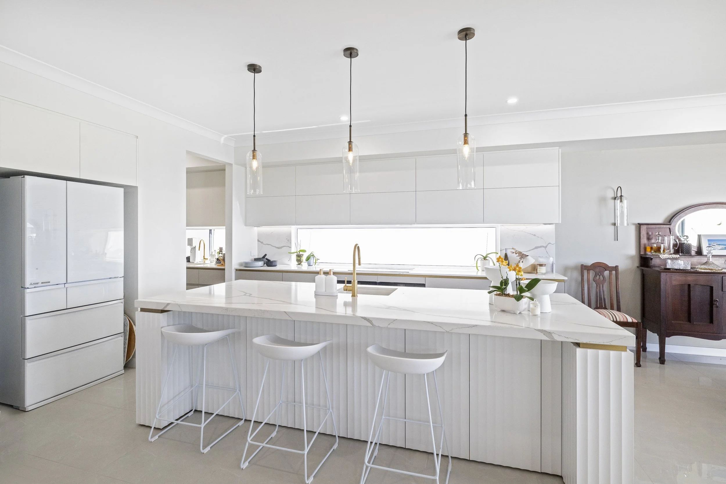 A modern, minimalist kitchen with white cabinetry, a white marble island with bar stools, pendant lights, and decorative plants.