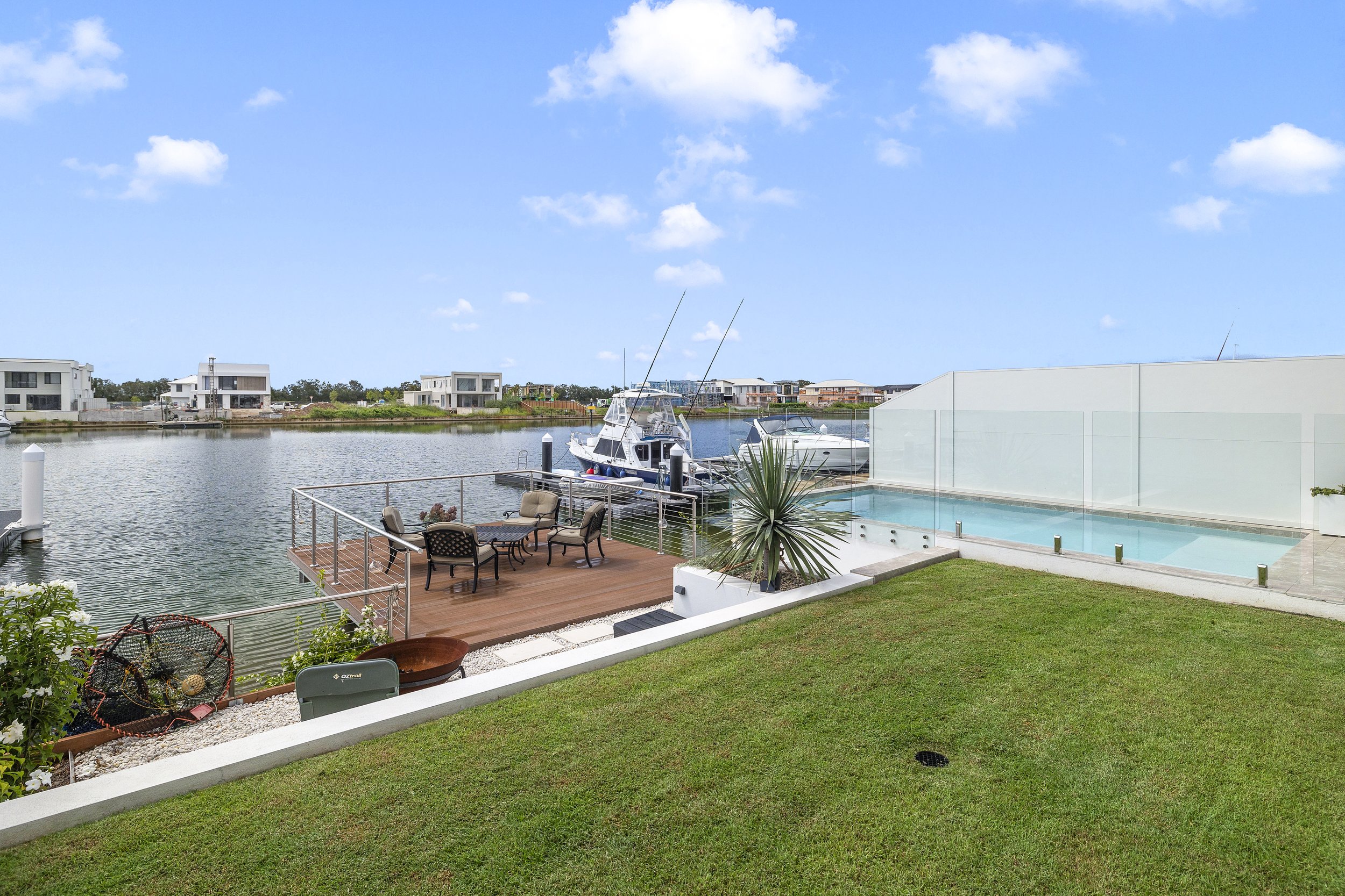 View of a modern house with a balcony overlooking a waterway. There are boats docked at the marina, a swimming pool, outdoor seating, and green grass area. In the background, several contemporary houses are visible on the opposite shore under a clear
